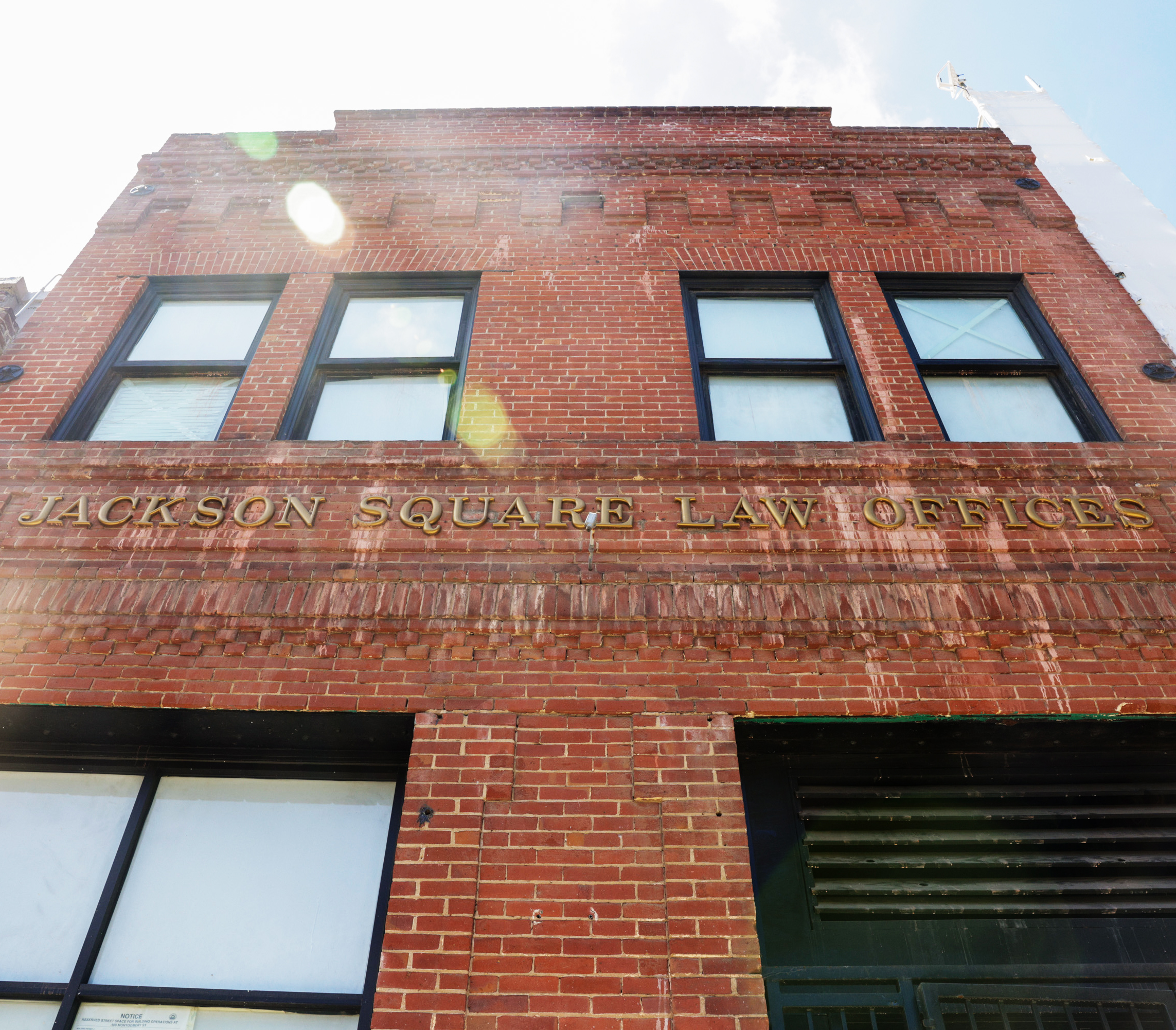A brick building with &quot;JACKSON SQUARE LAW OFFICES&quot; on the facade, featuring two windows and lens flare from the sun.
