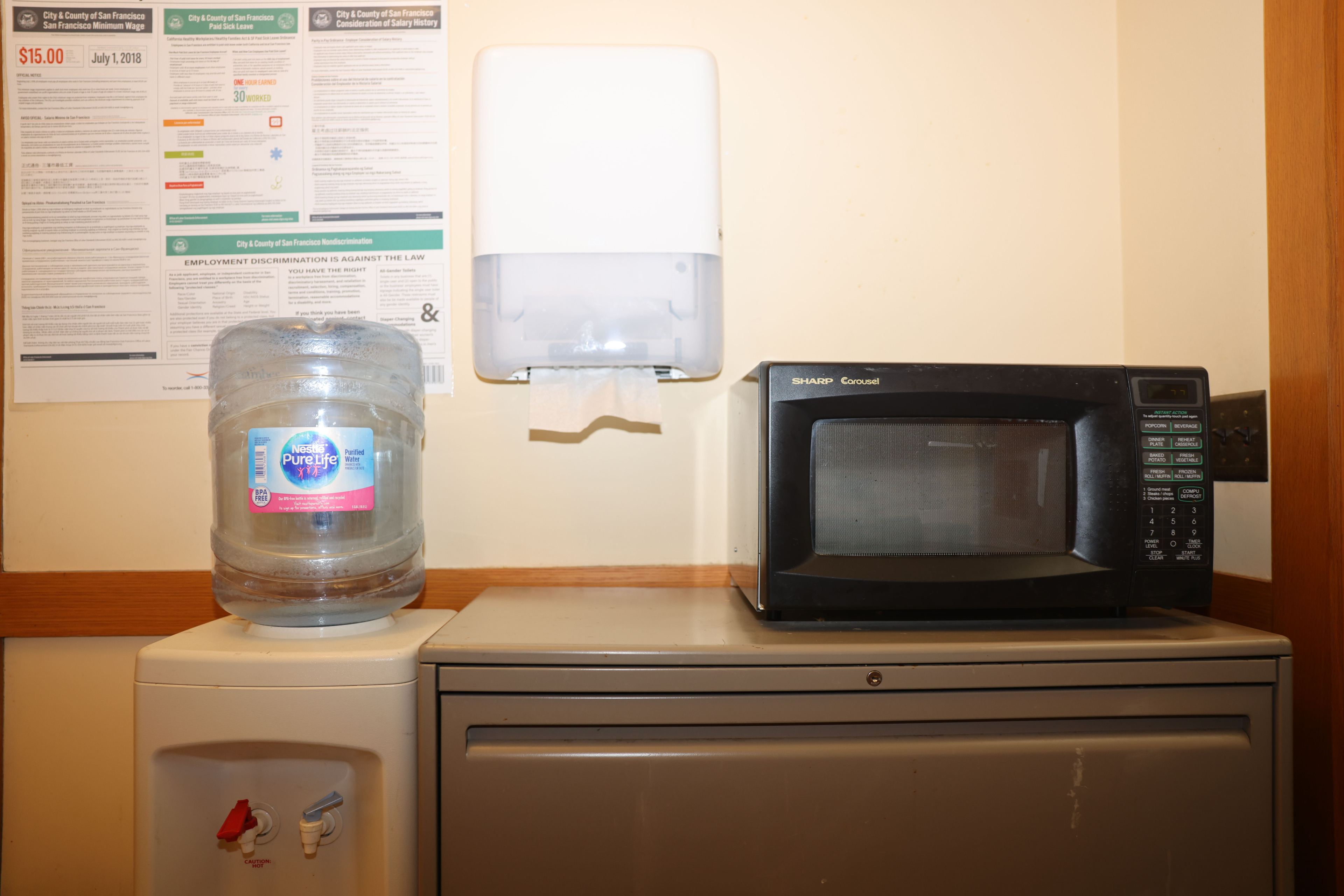 A water cooler, paper towel dispenser, microwave, and notices on a wall in an office break room.