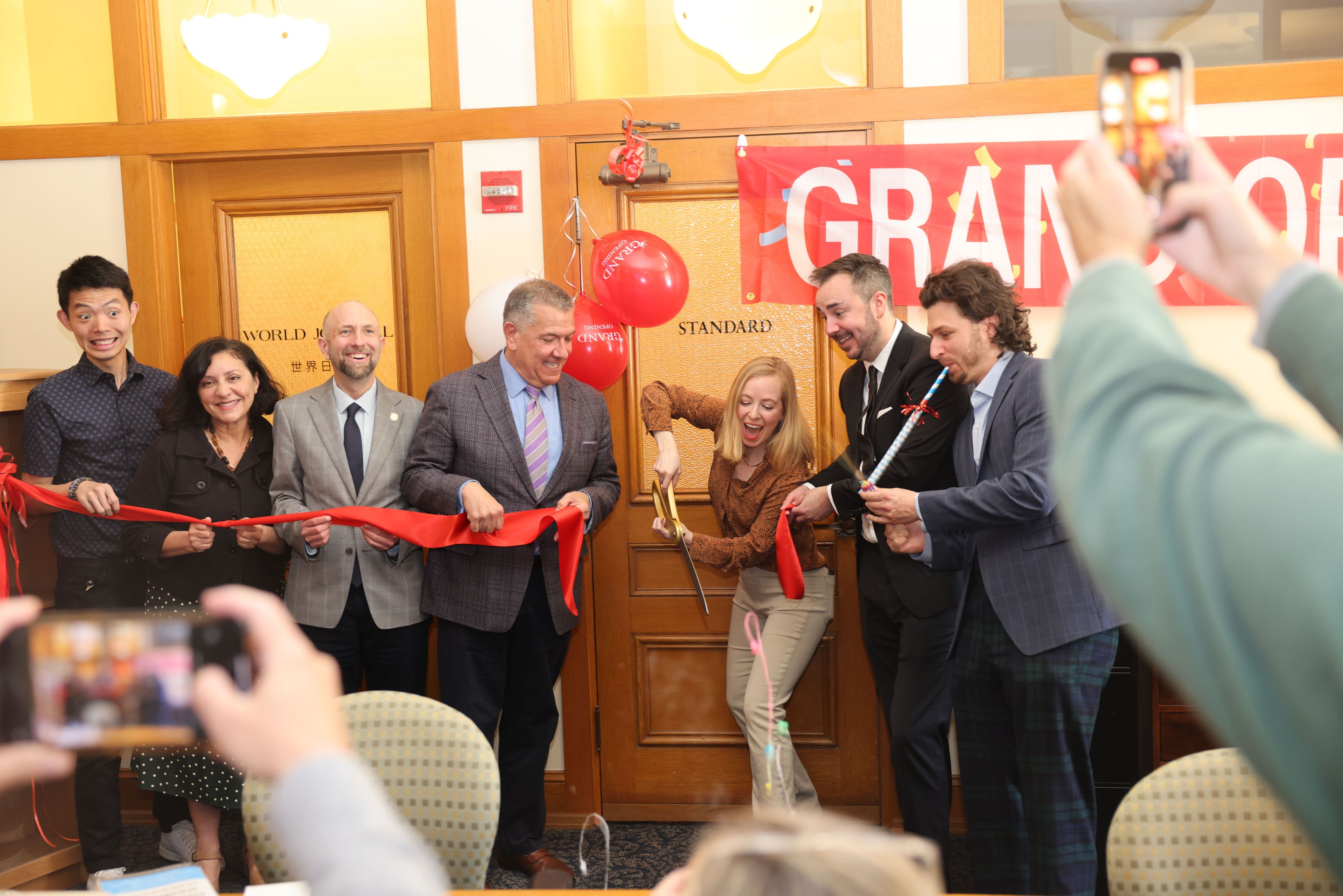A group is cutting a red ribbon at a grand opening event, smiling and posing for photos.