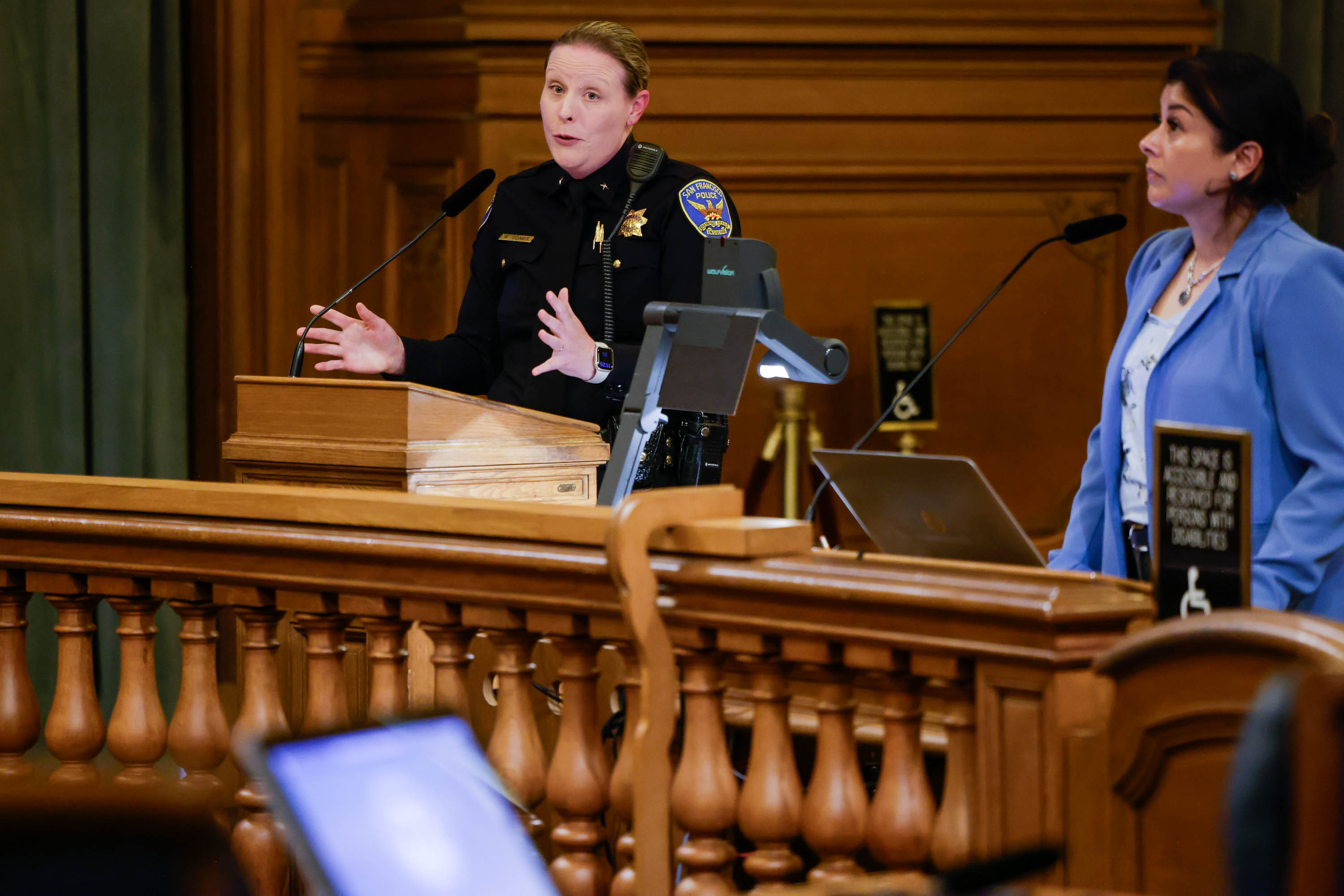 A uniformed police officer speaks at a podium while another person listens nearby in a wood-paneled room.