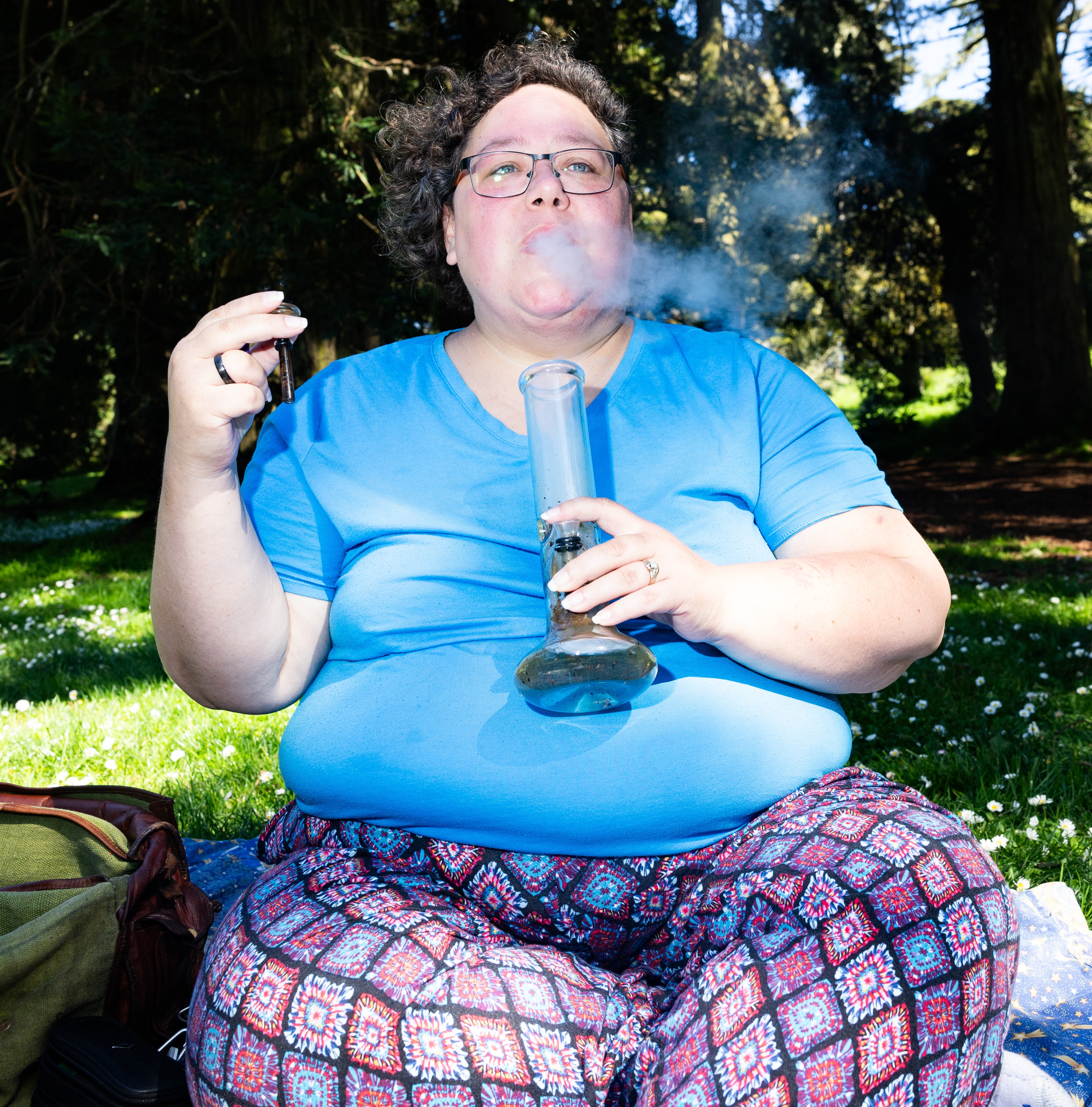 A person is sitting outdoors, inhaling from a bong with smoke visible, and holding a lighter.
