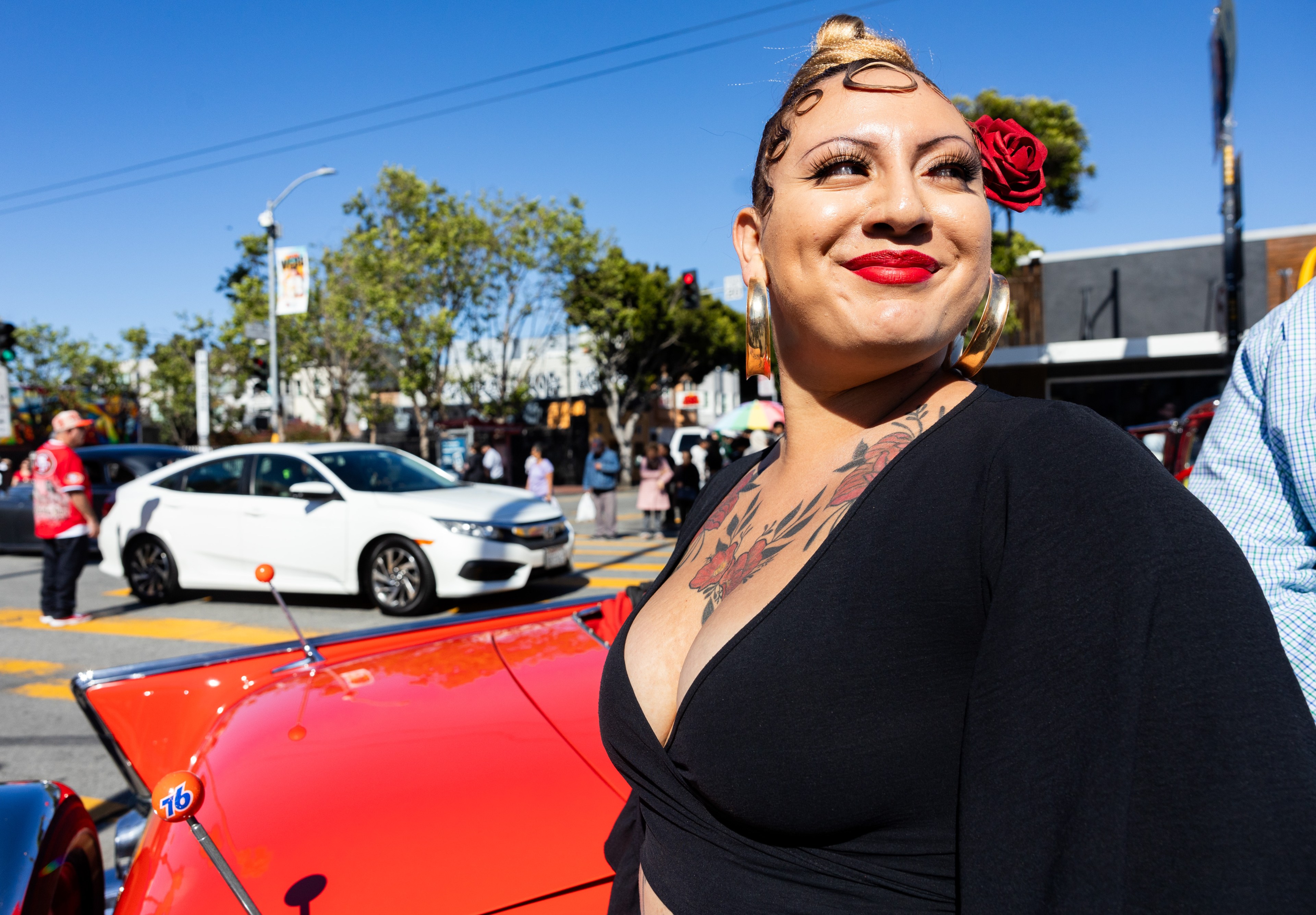 A woman with a red flower in her hair smiles on a sunny street with cars and pedestrians in the background.