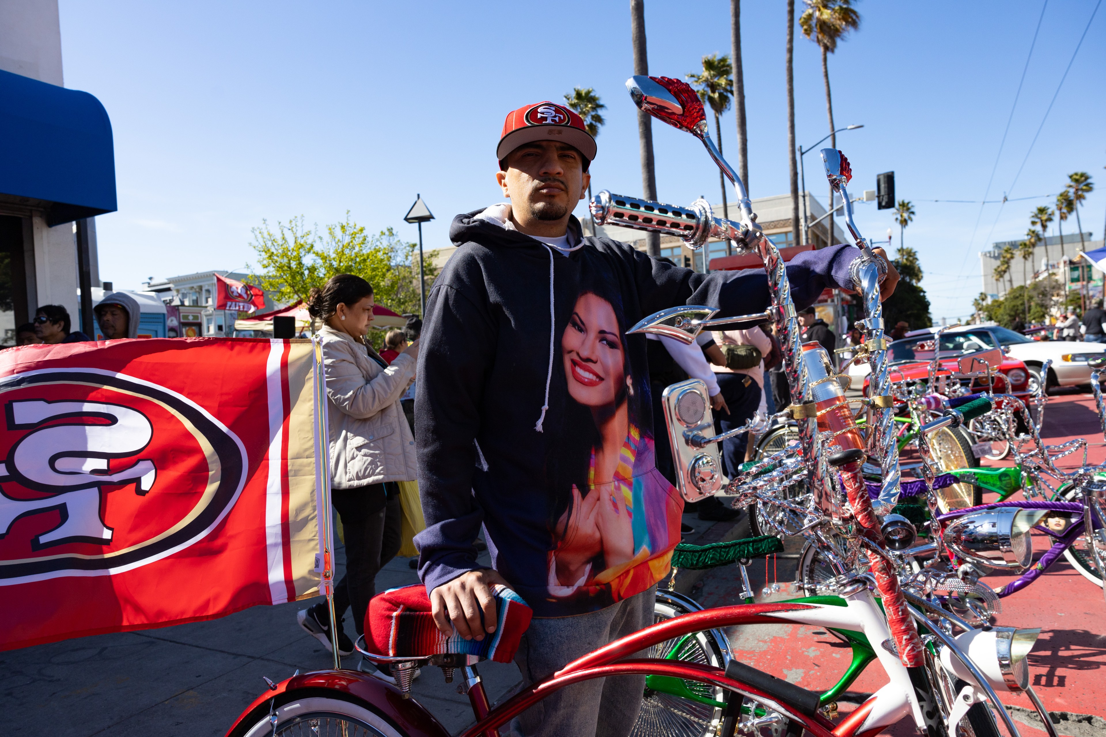 A man stands with custom lowrider bicycles, wearing a cap and a hoodie with a woman's face. A sports team flag is in the background.