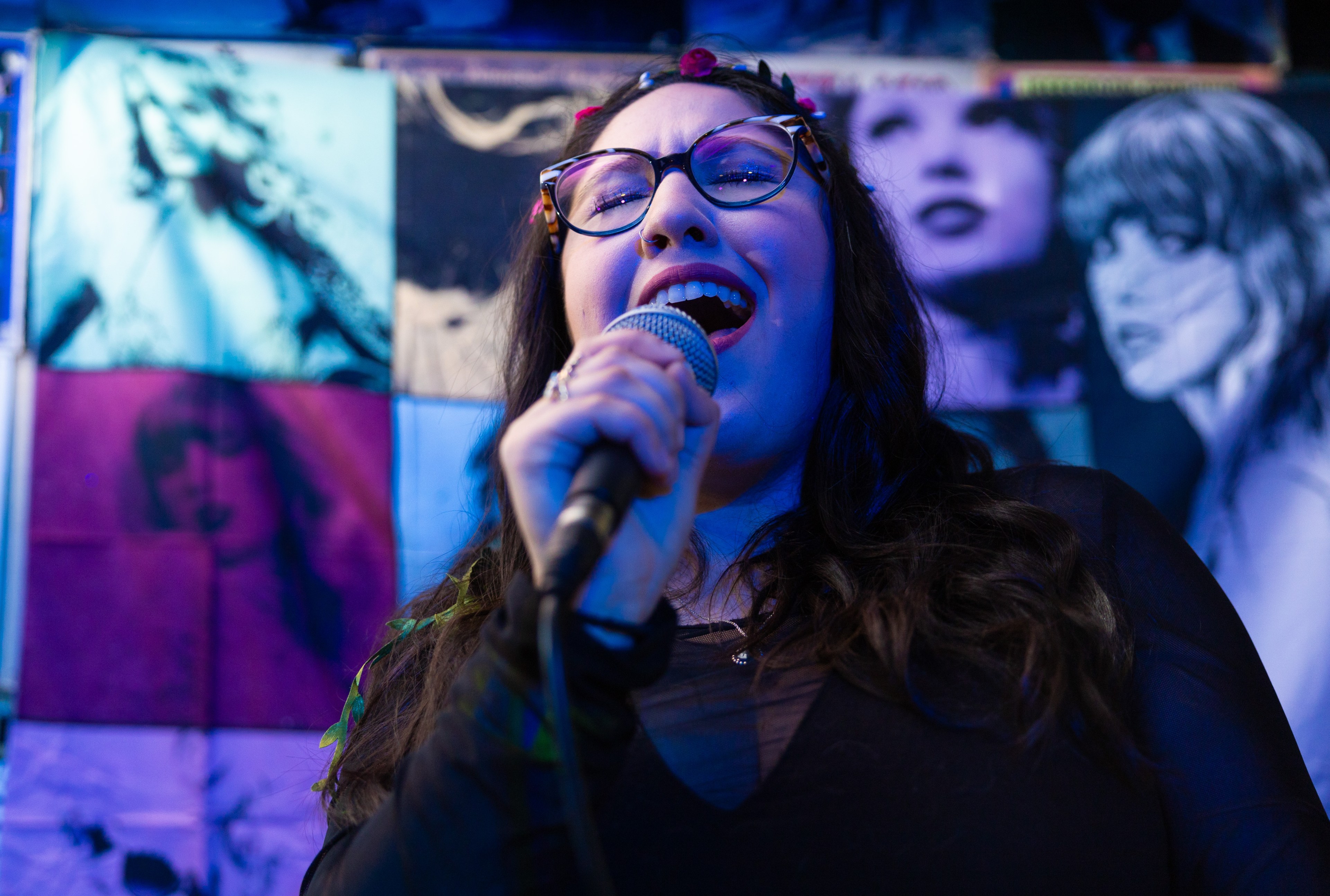 A woman sings into a microphone with vibrant wall art of female faces behind her.