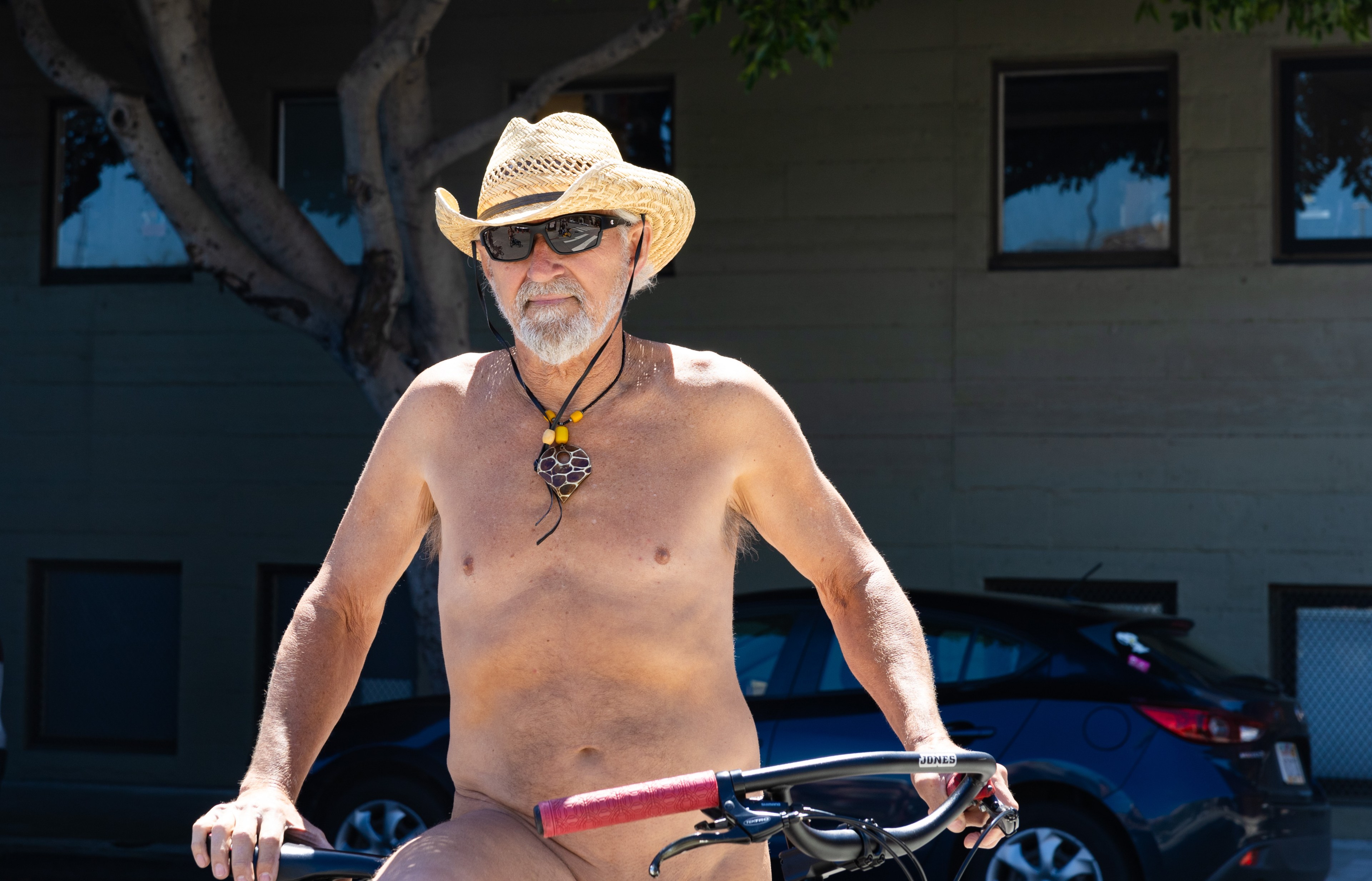 A person with a straw hat and sunglasses is sitting on a bike, shirtless, with a building and a car in the background.
