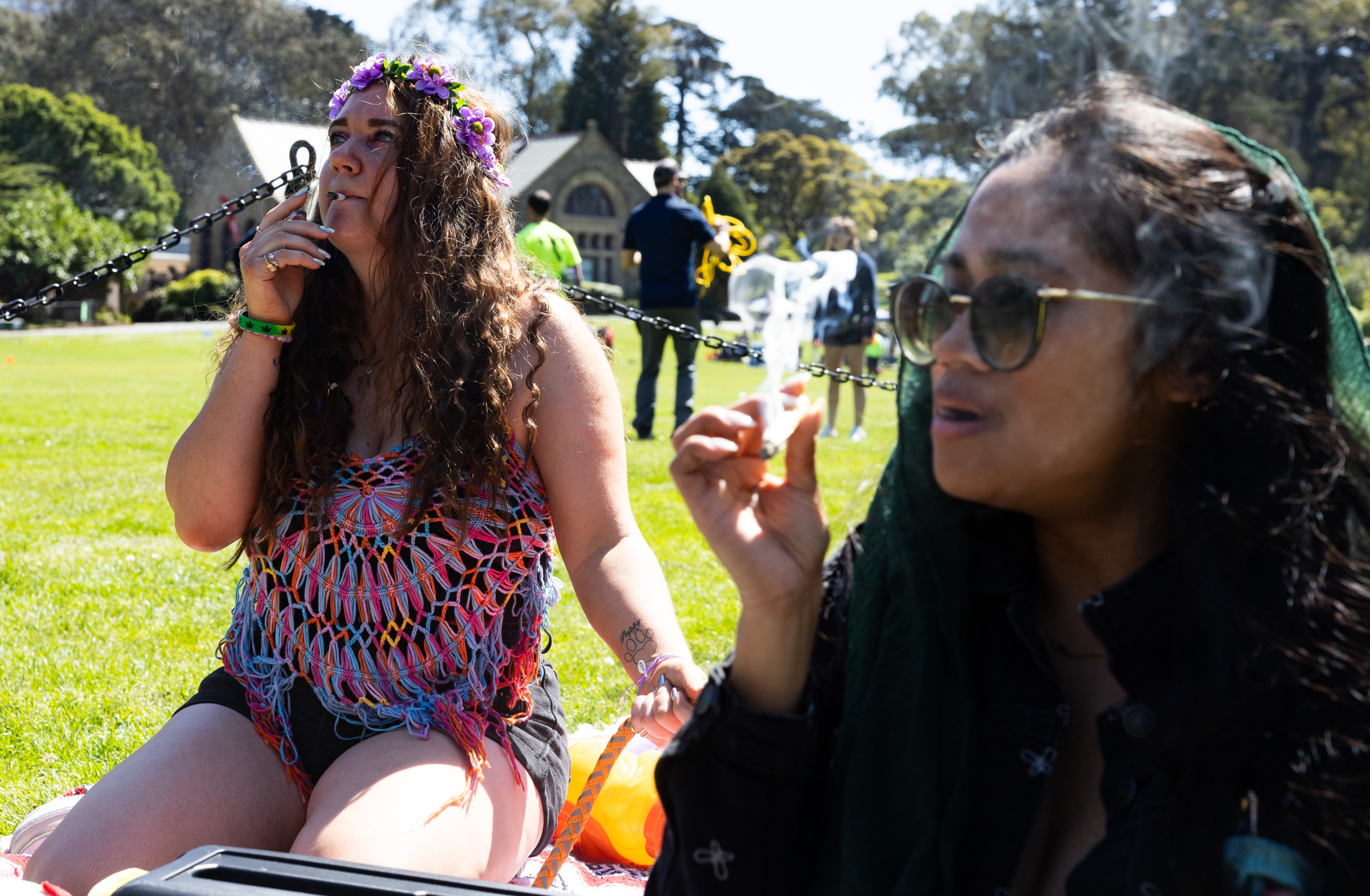 Two women sit on a sunny park lawn; one wears a flower crown and smokes, the other holds sunglasses and talks.