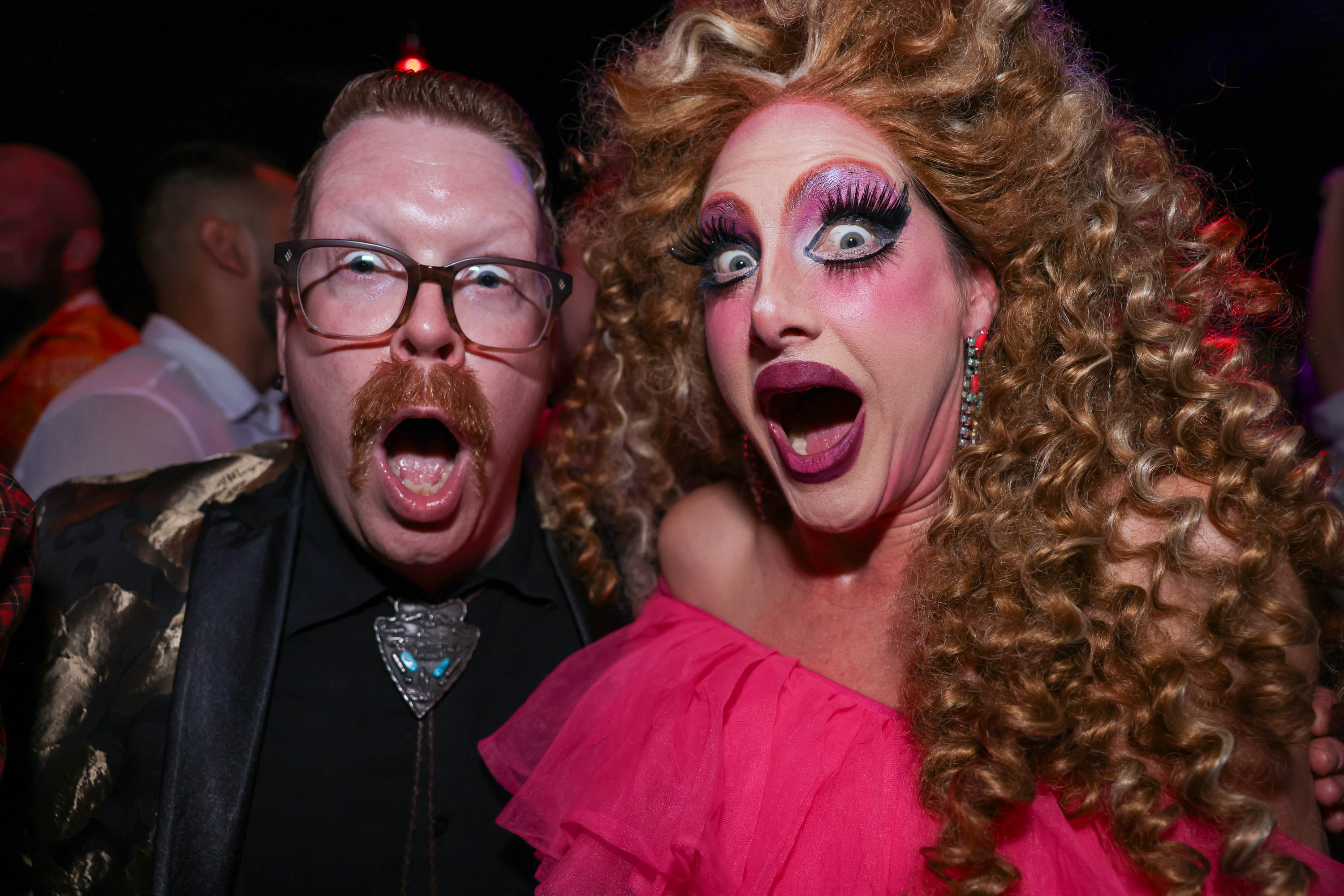 two white drag performers open their mouths wide in mock gasps, one with a mustache and glasses, the other with huge eye makeup and a curly blond wig