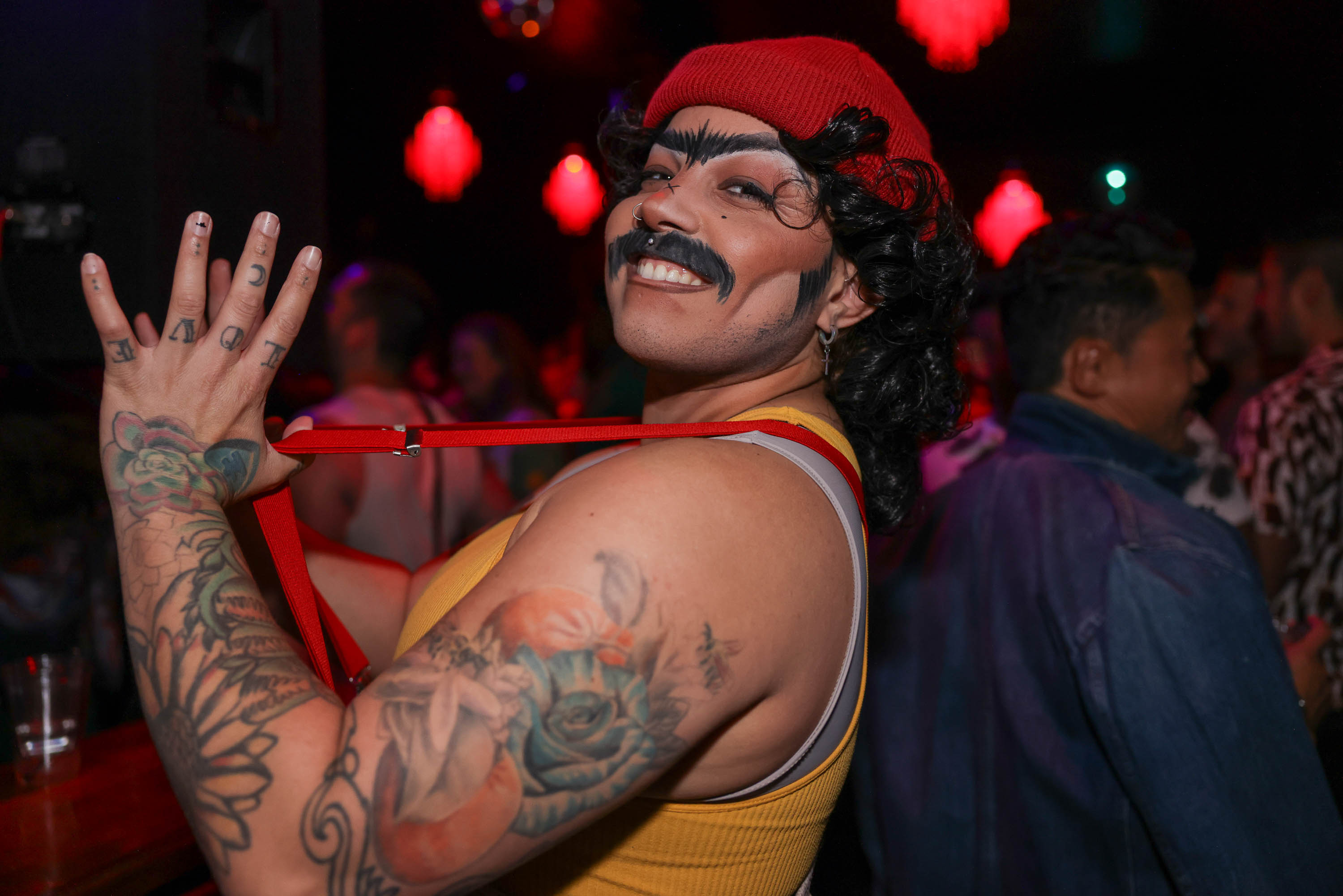 a smiling, mustachioed person in a red hat flicks a red suspender, their tattooed arm exposed
