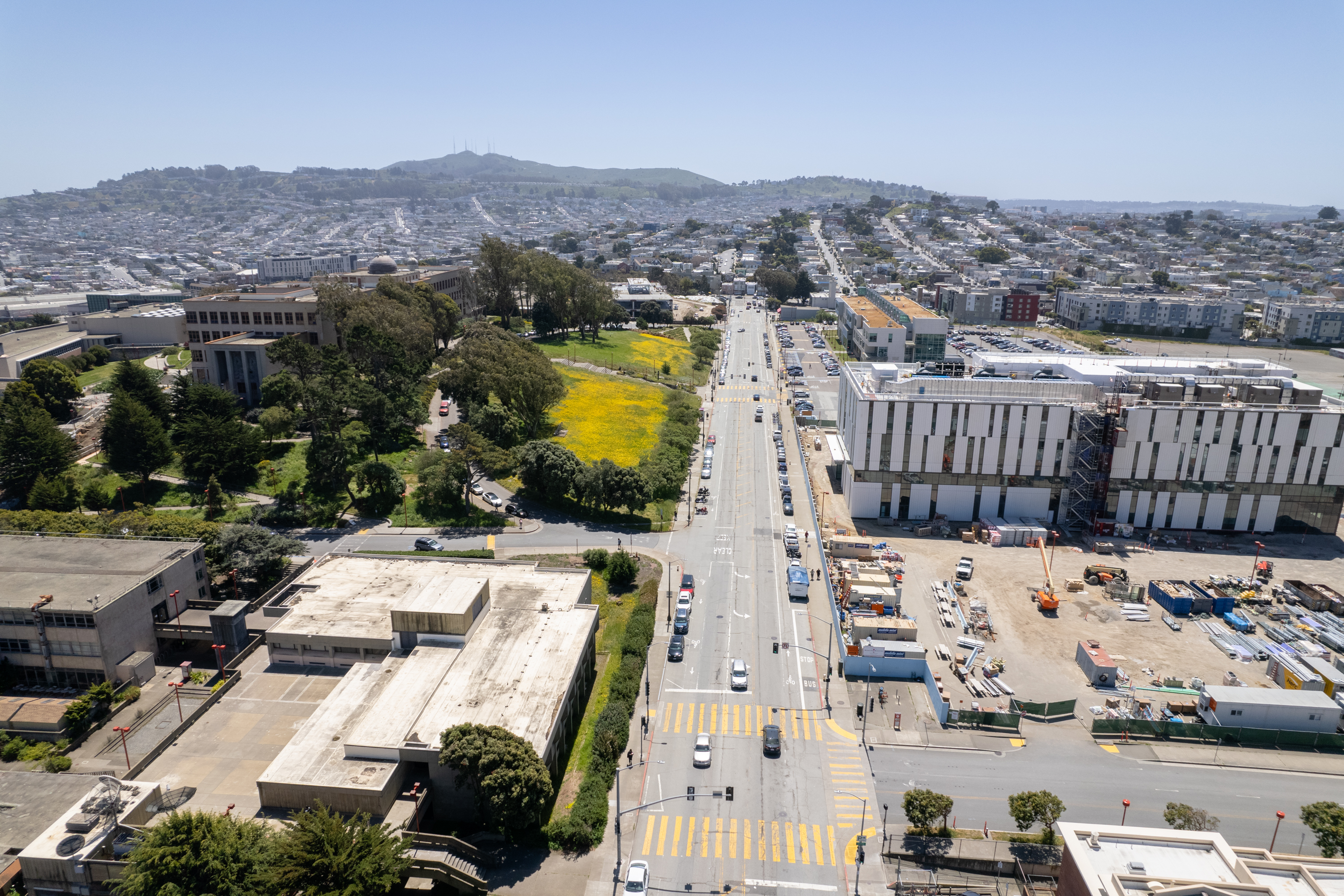 Aerial view of a city street with buildings, construction, green spaces, and distant hills on a clear day.