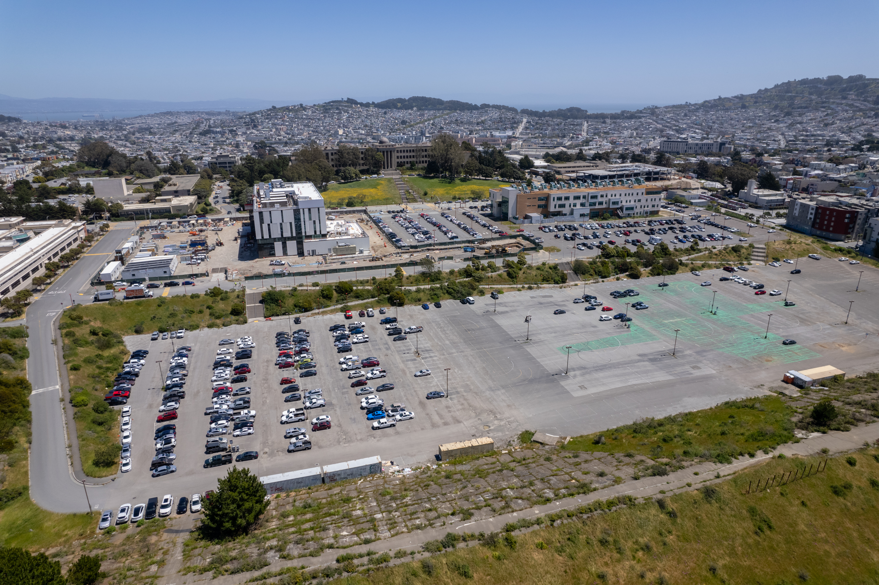 An aerial view of a large parking lot with cars, buildings nearby, and a hilly cityscape in the distance.