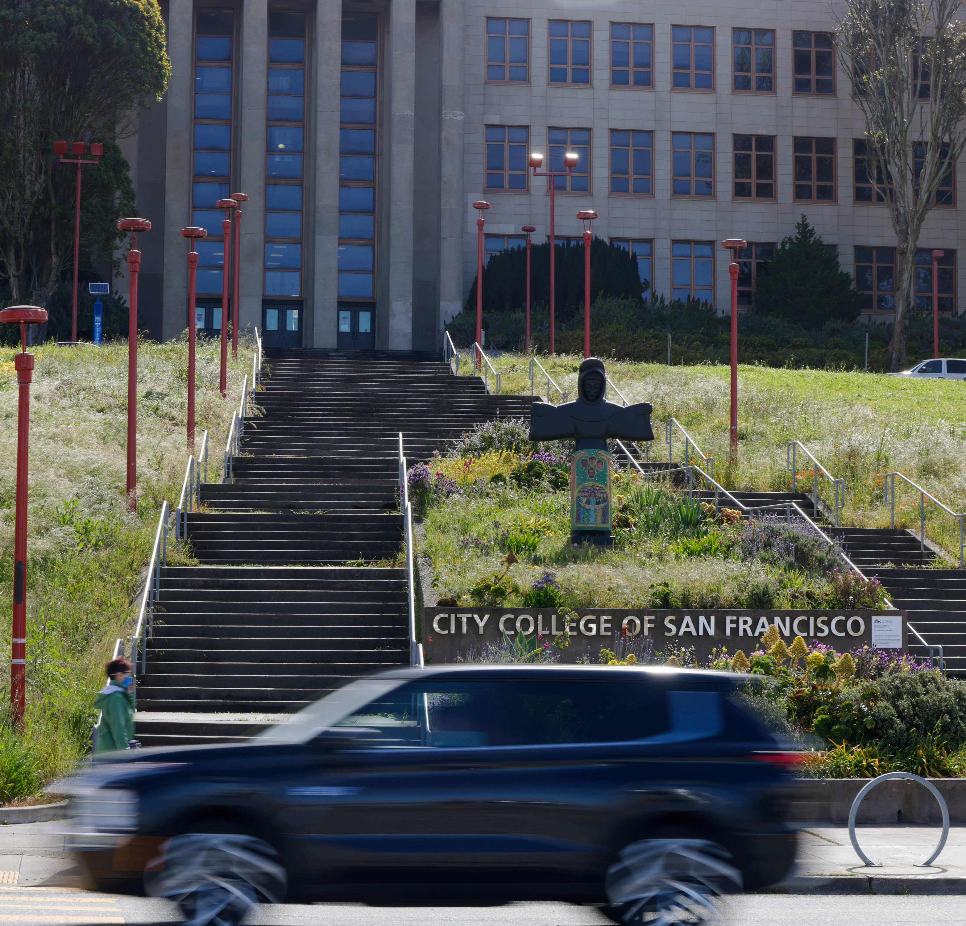 A car speeds past steps leading up to City College of San Francisco, flanked by red lamp posts and a colorful statue.