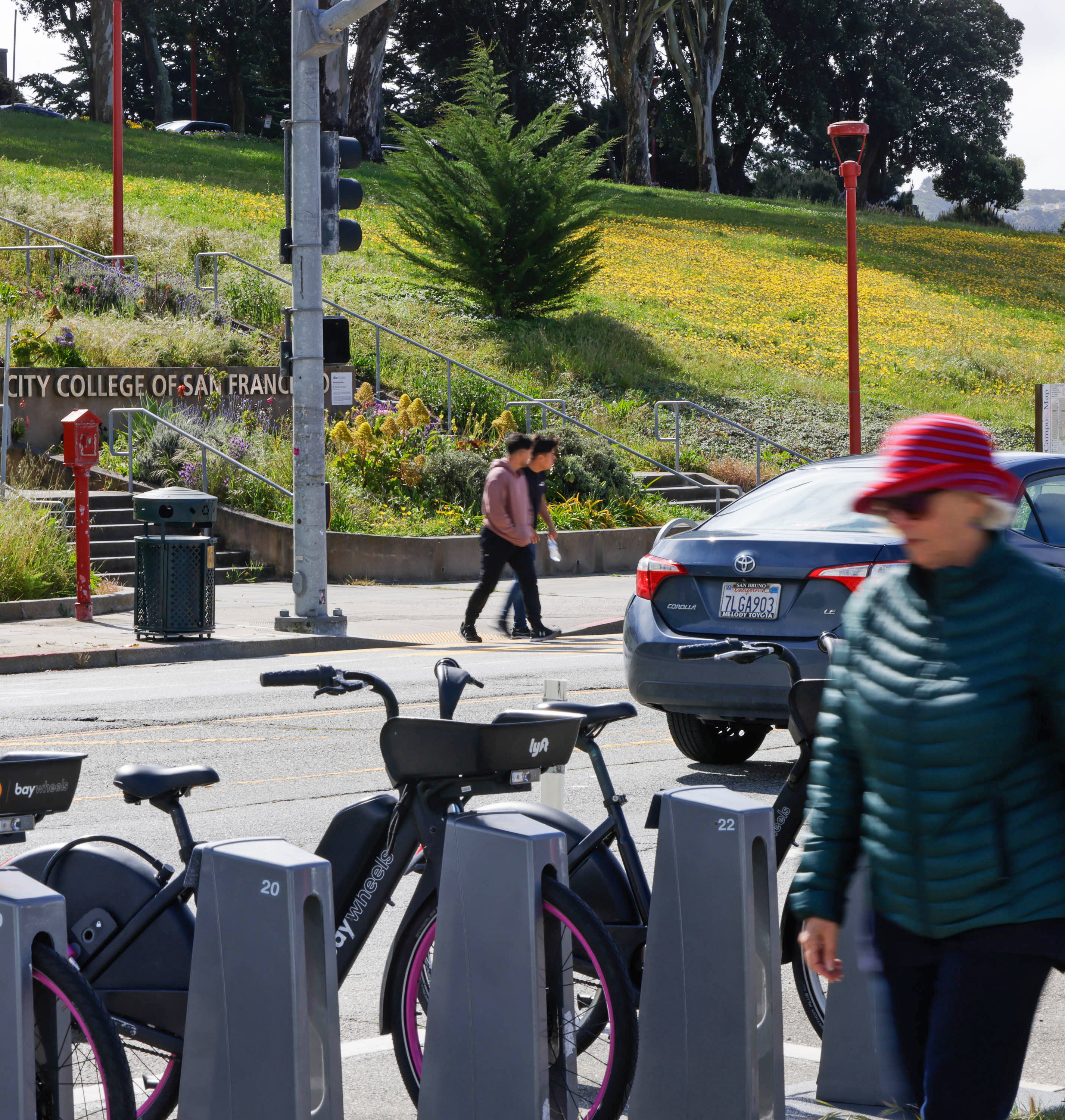 A busy sidewalk with a bike share station, people walking, a car, and a lush hillside with yellow flowers in the background.