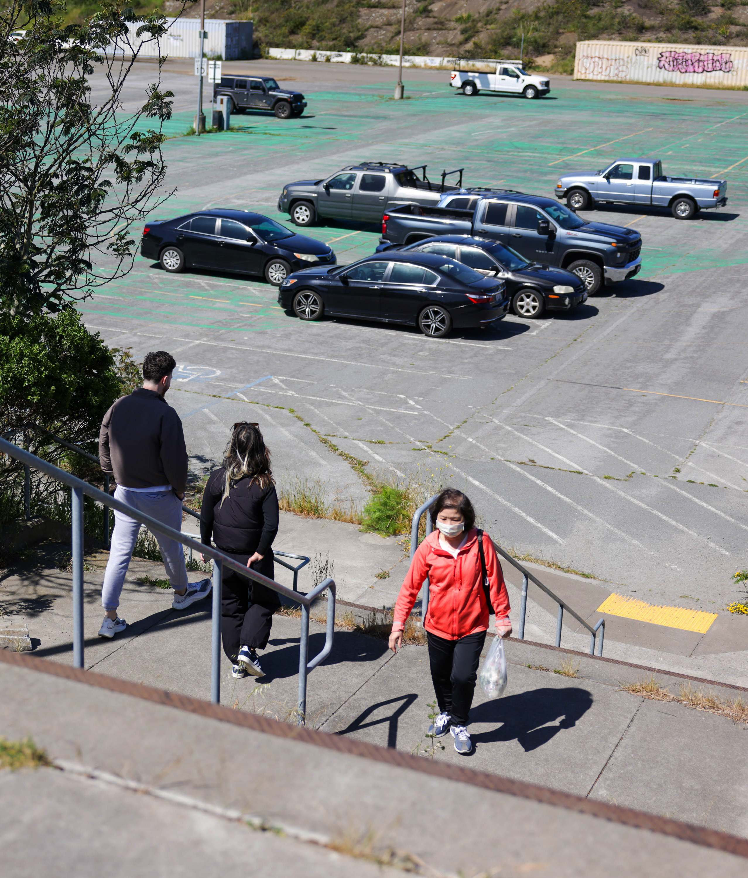 Three people are walking up concrete steps beside a parking lot with several cars.