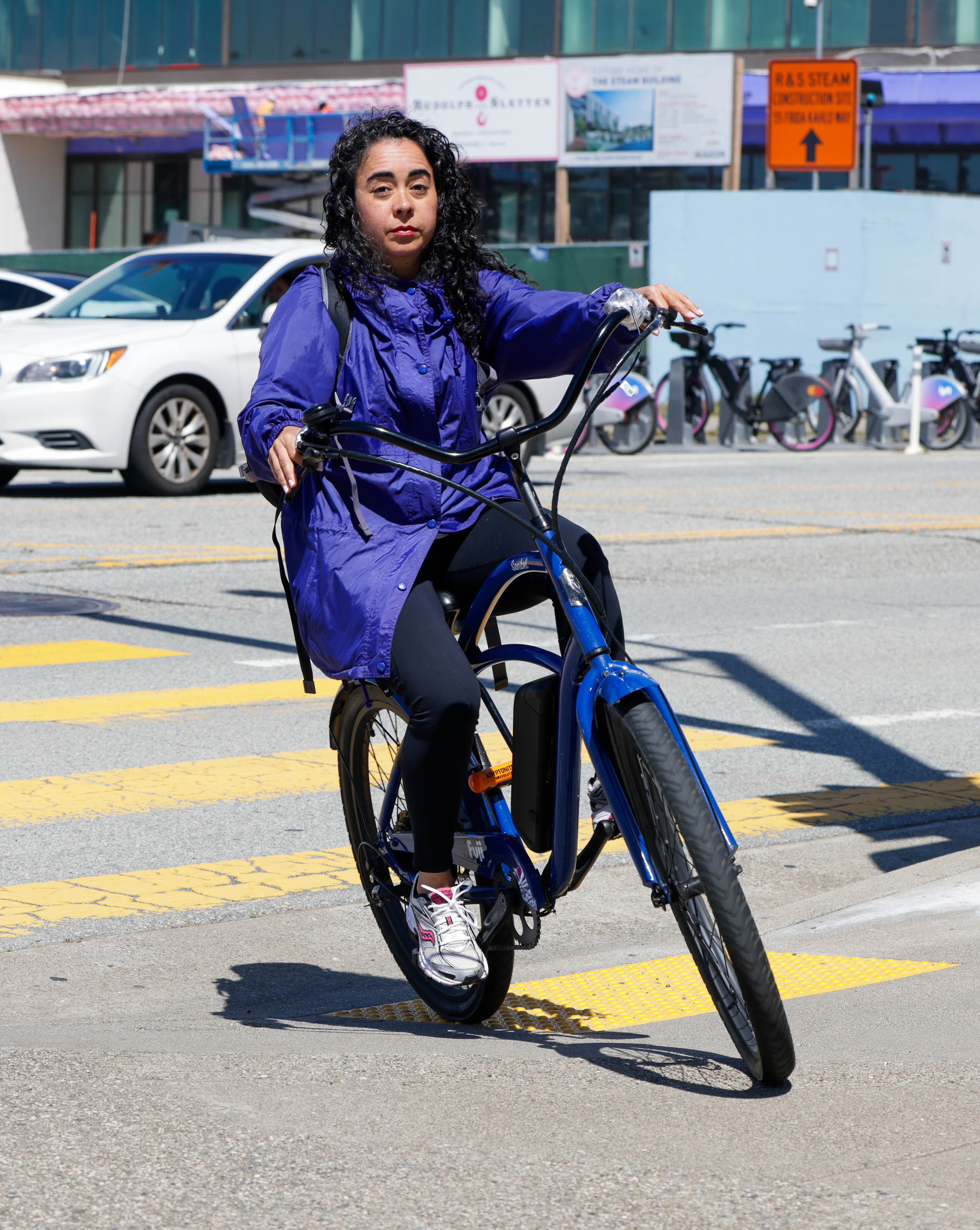 A person in a blue coat rides a blue bike across a crosswalk, with parked cars and buildings behind.