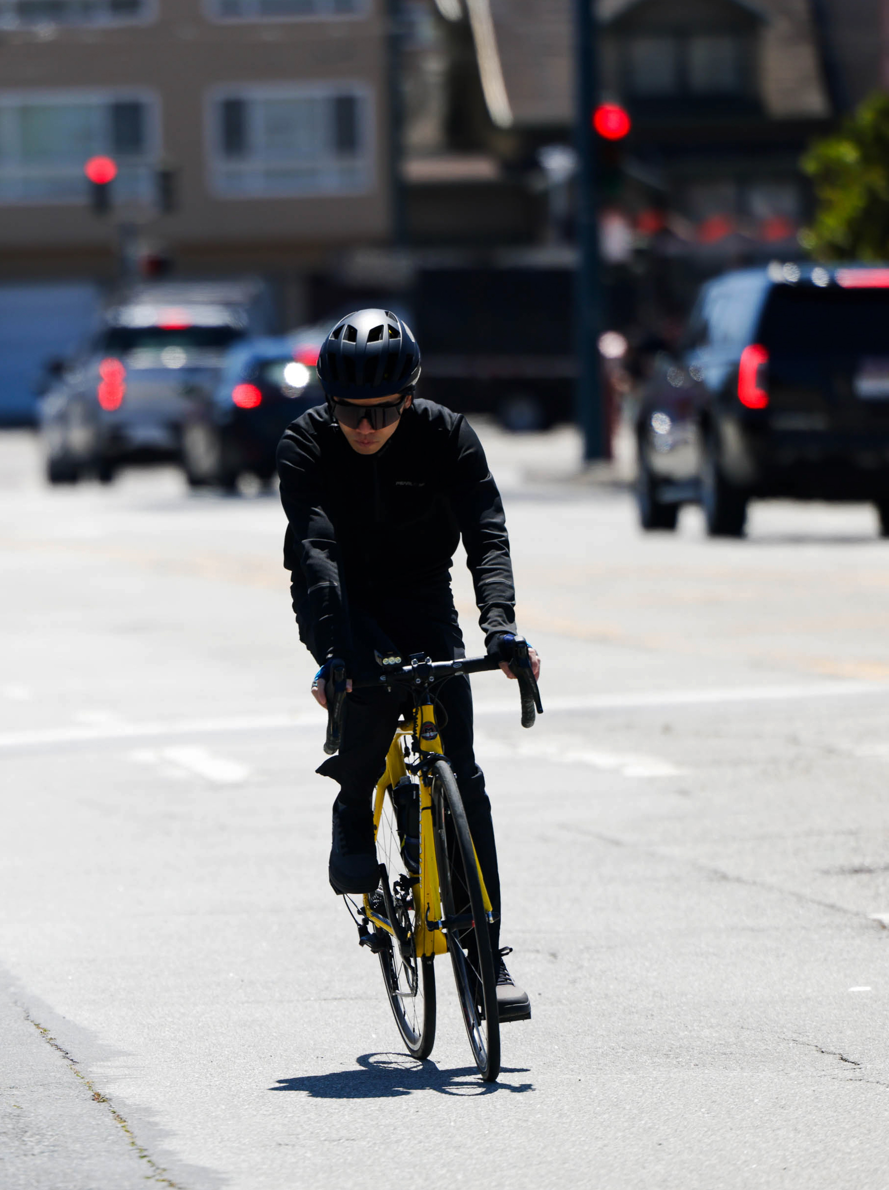 A cyclist in black attire rides a yellow bike on a sunny street, vehicles and traffic lights behind.