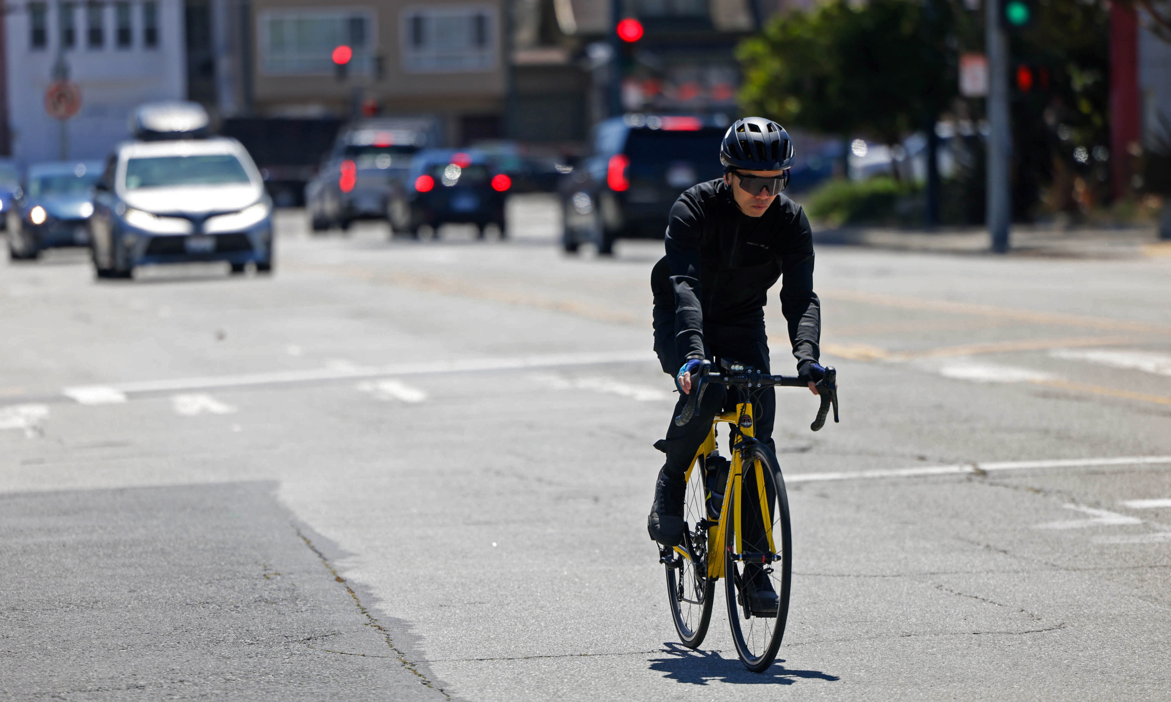 A cyclist in black attire and helmet rides a yellow bike on a sunny city street with cars and traffic lights.