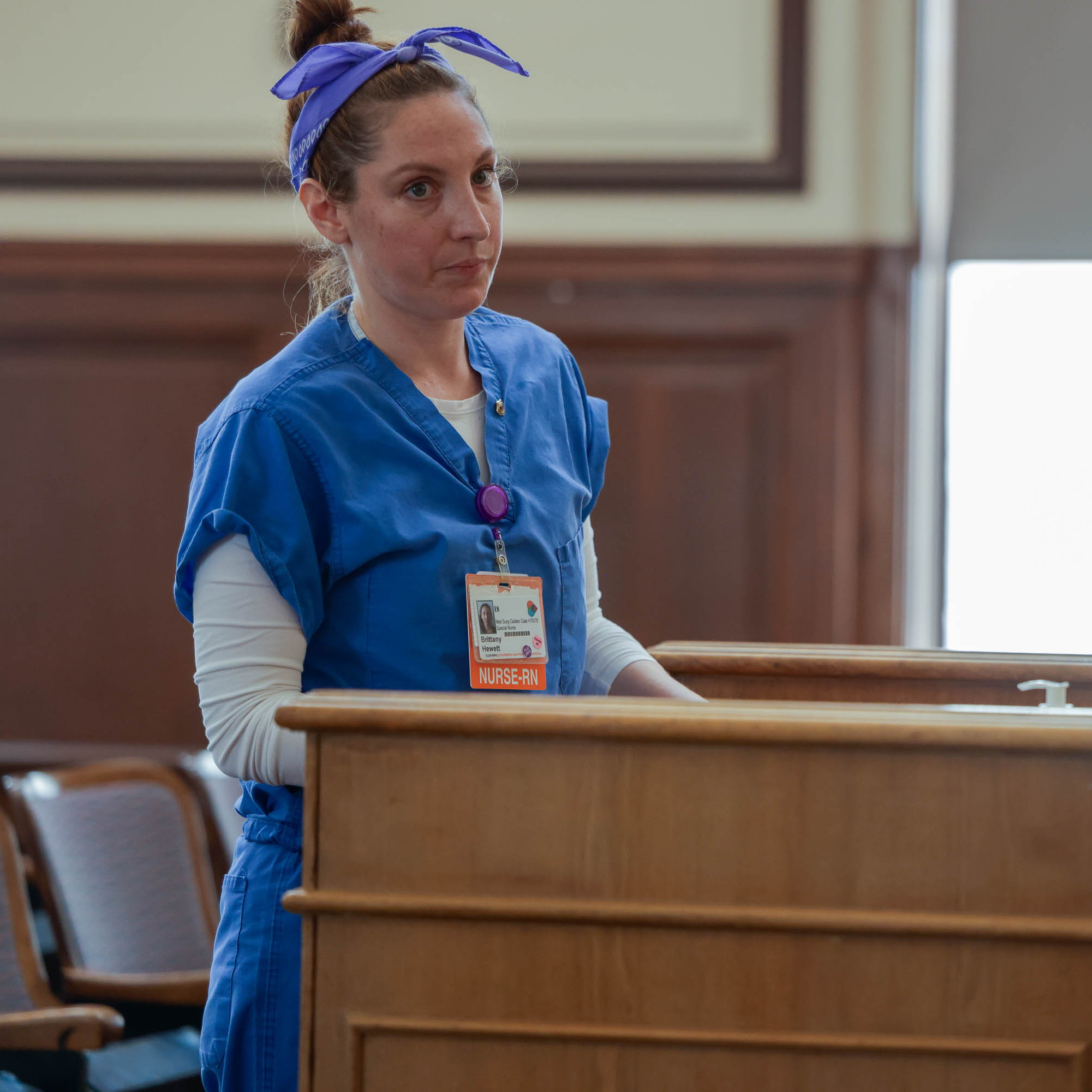 A nurse in blue scrubs stands by a wooden bench, her name badge visible.