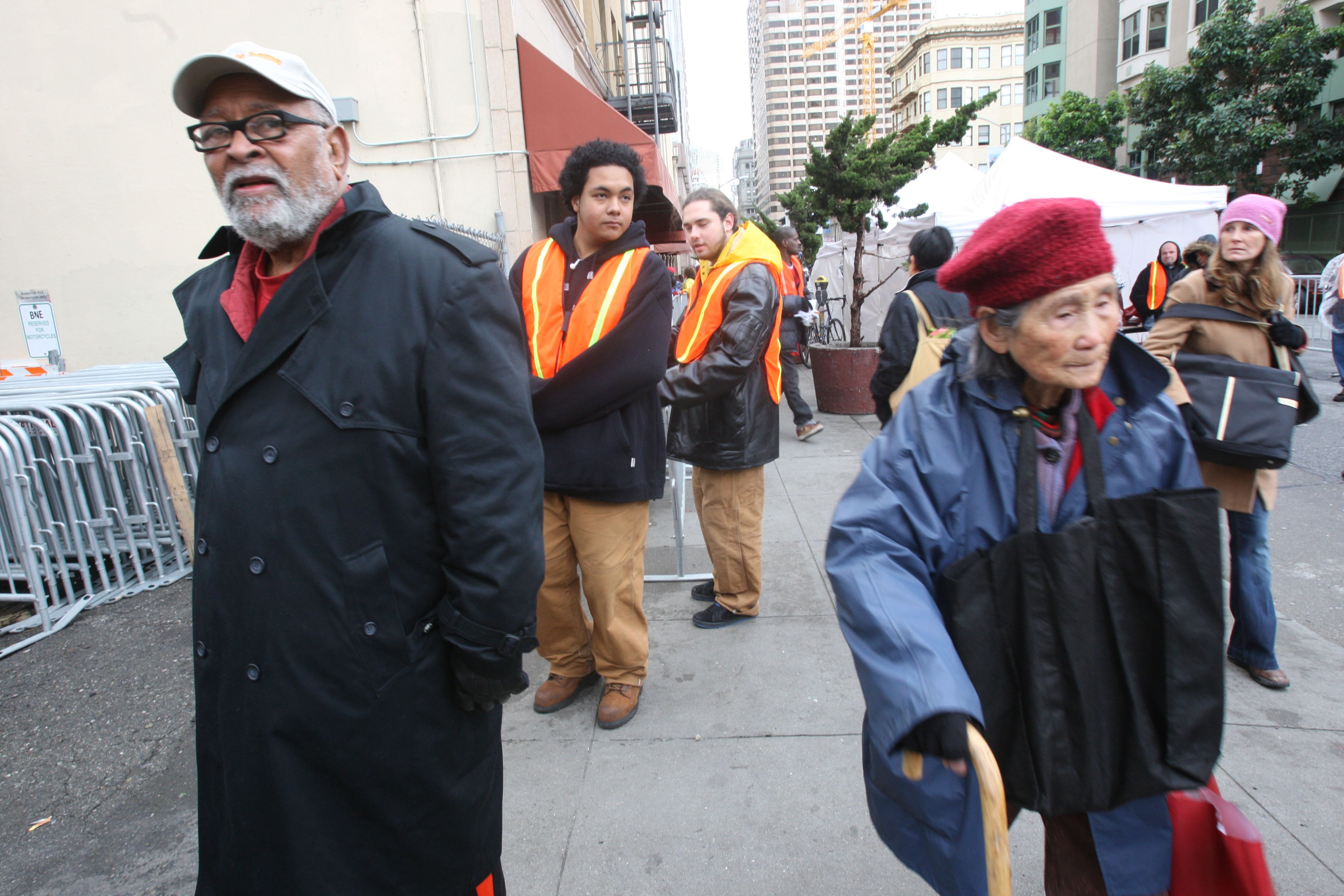 An elderly lady using a cane walks past various onlookers, including two men in high-visibility vests.