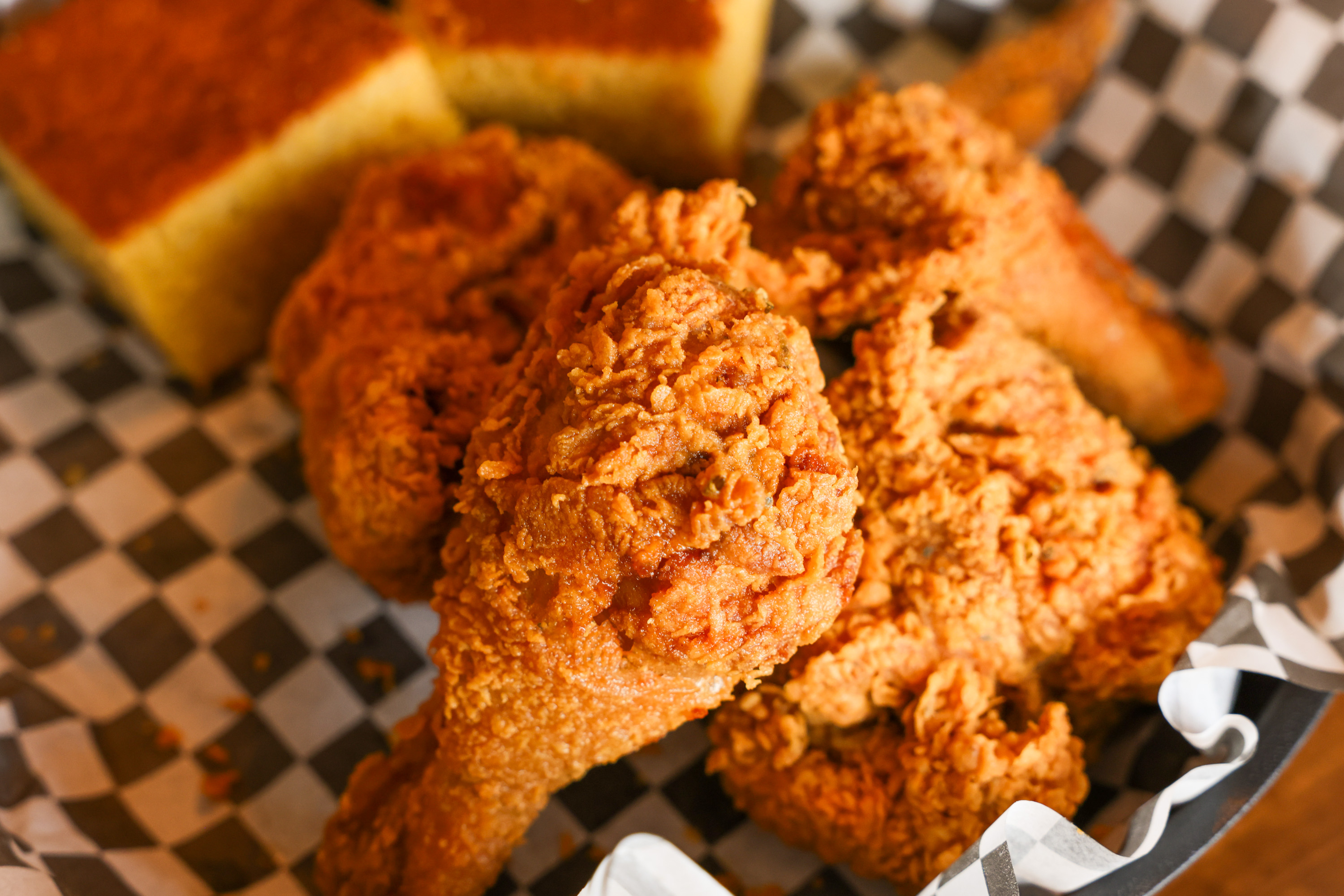 Crispy fried chicken pieces beside cornbread in a basket with checkered paper liner.