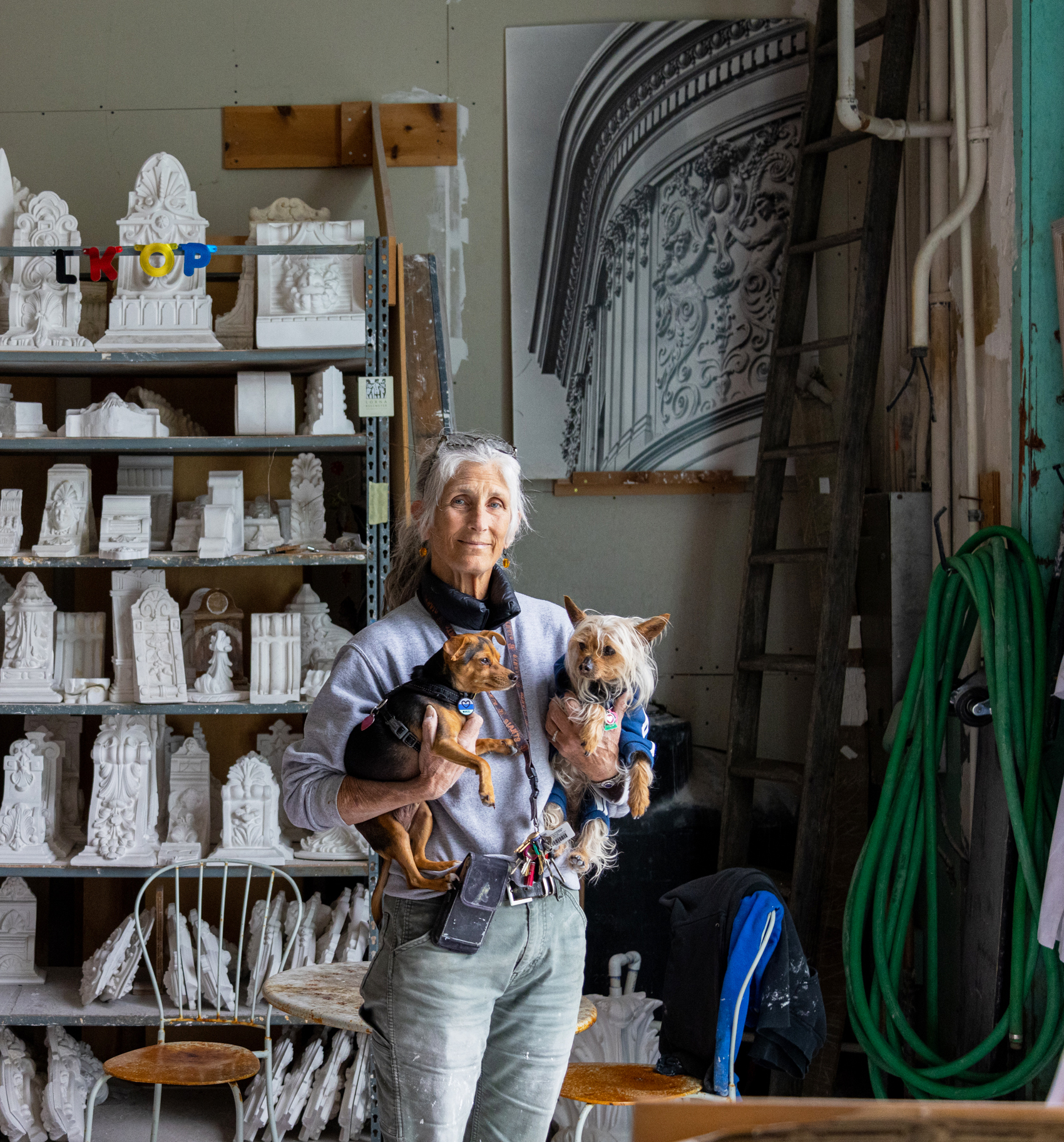 A woman holding two dogs stands in a workshop surrounded by plaster sculptures and molding tools.