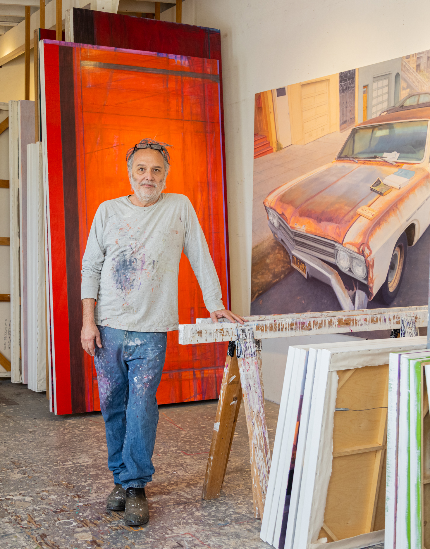 An artist stands in a studio with a large abstract red painting and a realistic car painting. He wears splattered clothes.