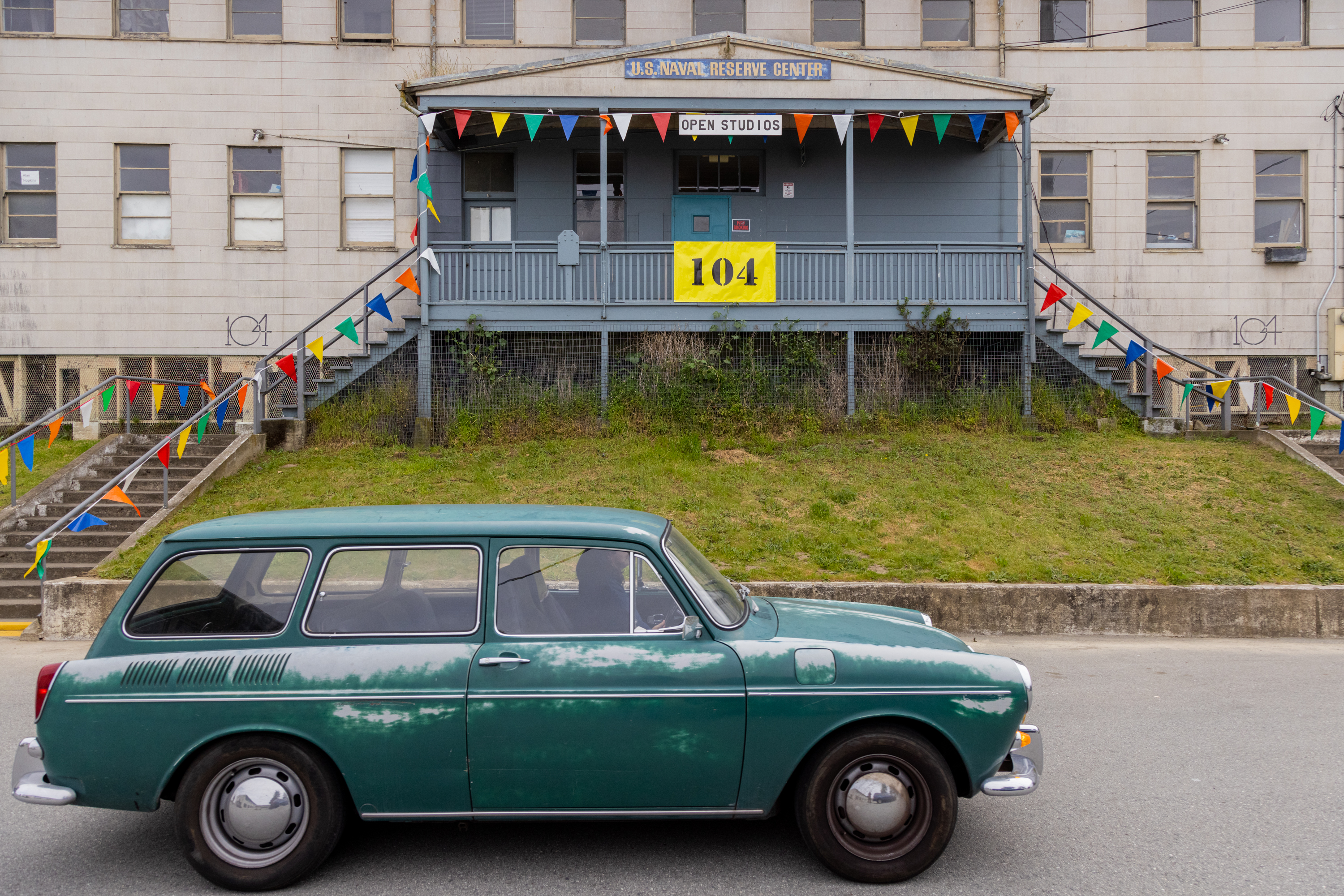 A vintage green car parked in front of a building with colorful banners and a sign &quot;OPEN STUDIOS&quot;.