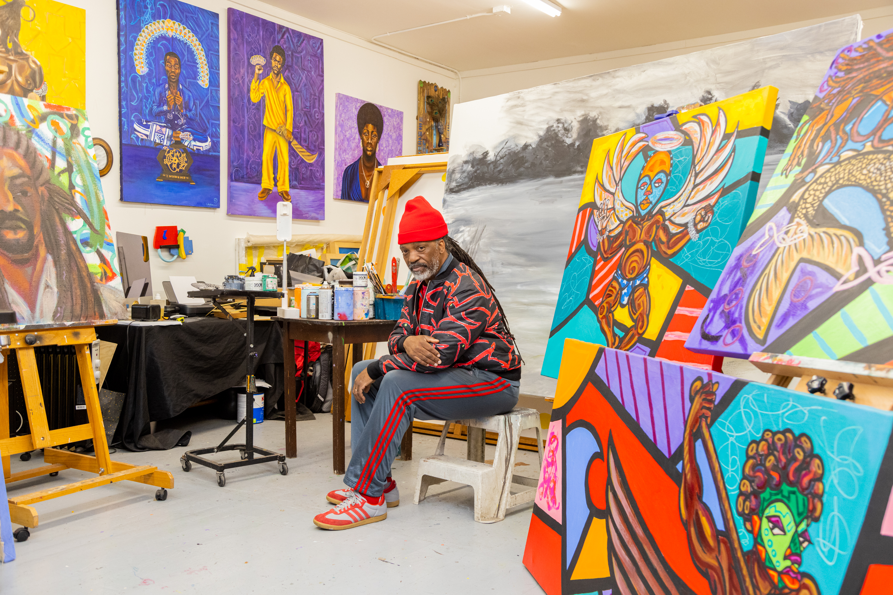 An artist sits in a studio surrounded by colorful, vibrant paintings on the walls and easels.