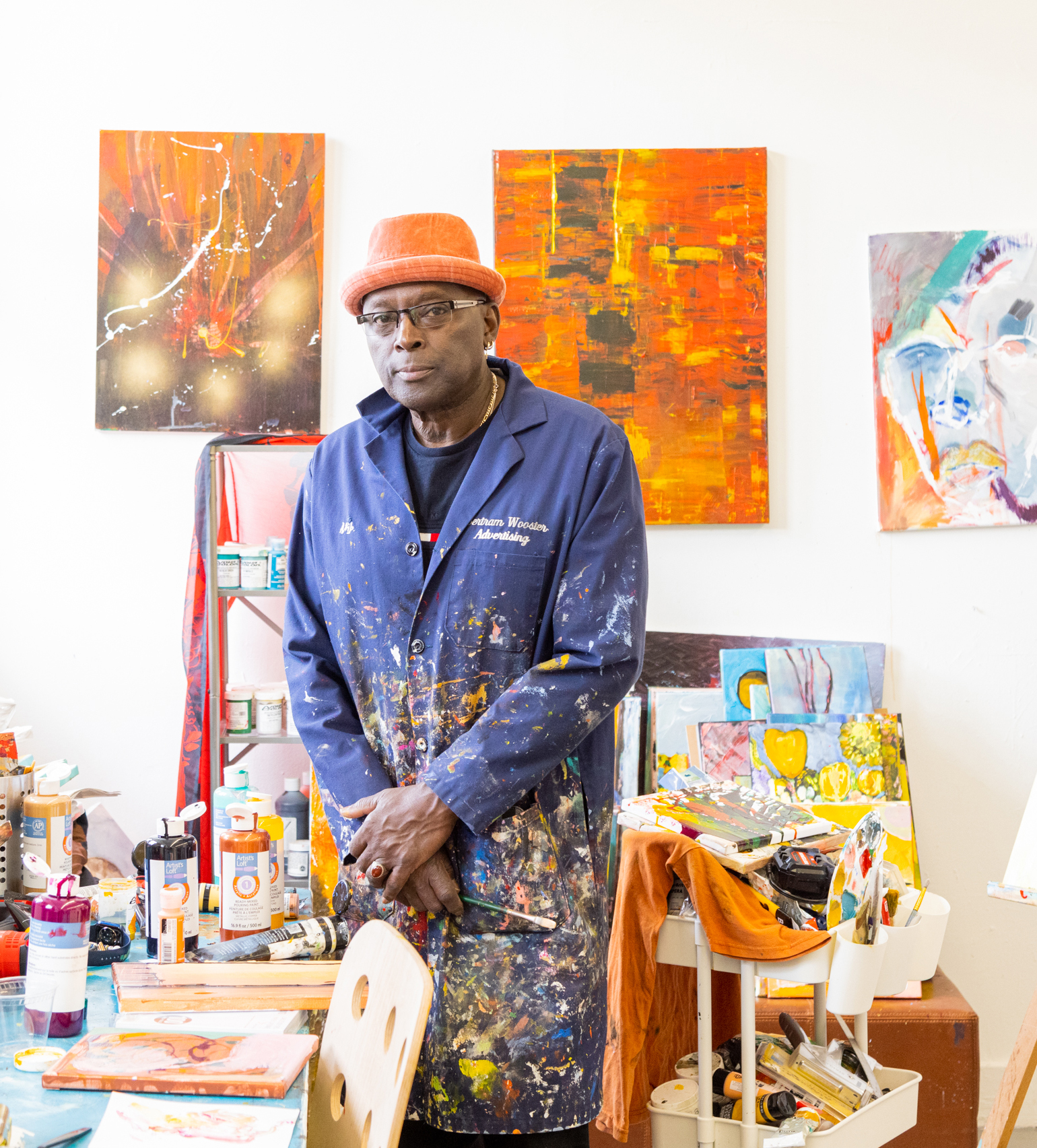 An artist in a paint-splattered coat stands in a studio surrounded by vibrant paintings and art supplies.