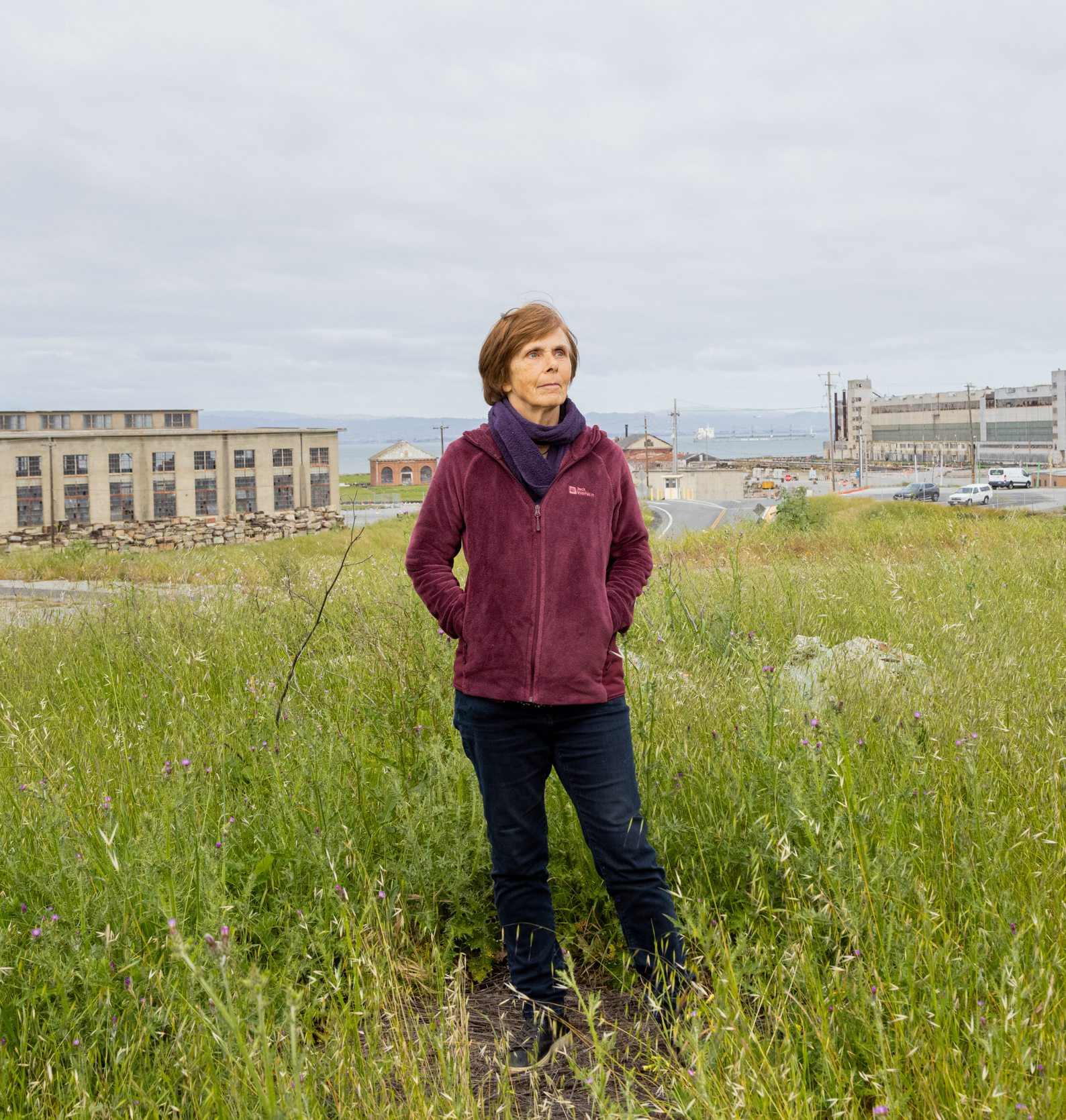 A person stands in a green field with industrial buildings in the background, under an overcast sky.