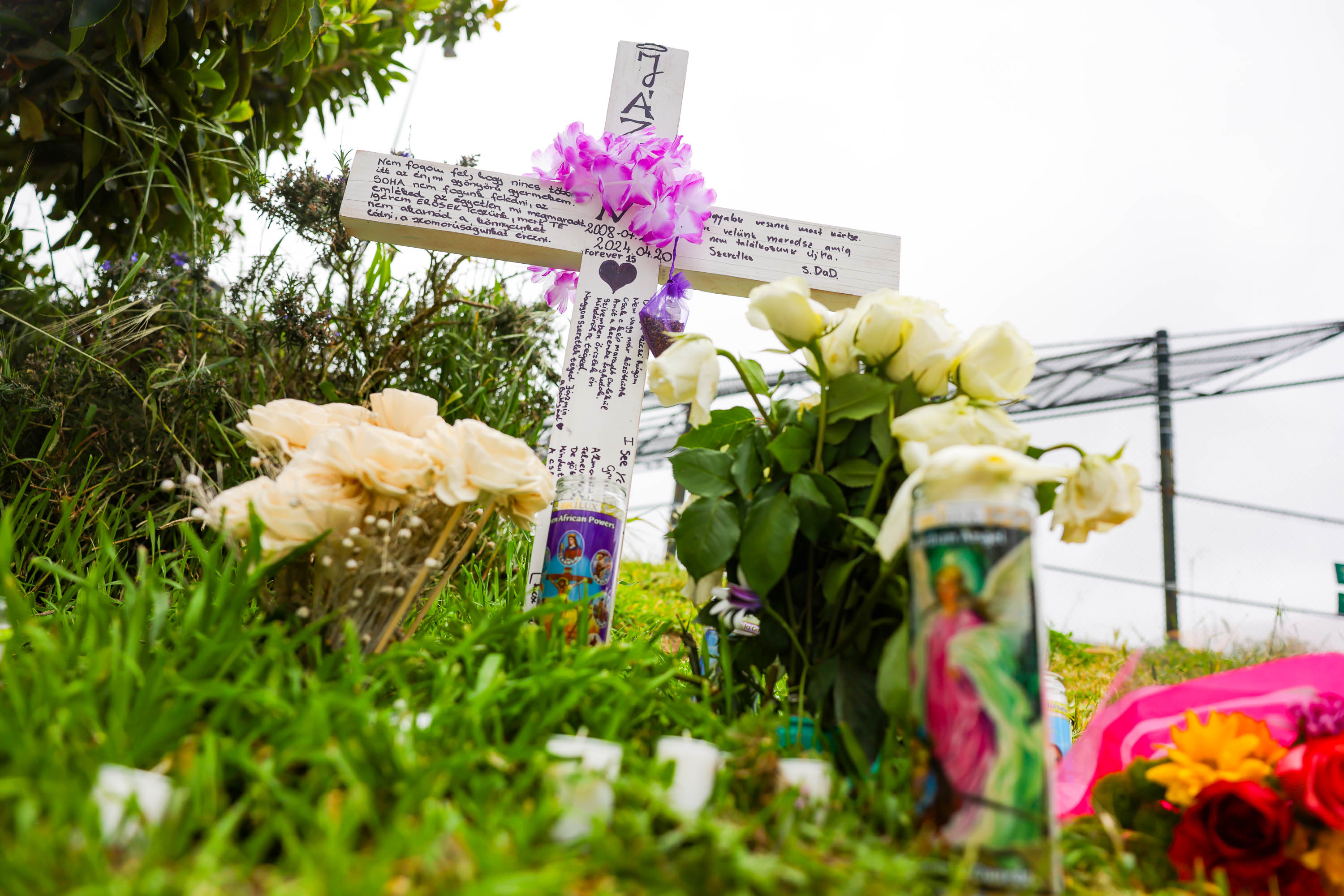 A makeshift roadside memorial featuring a white cross with flowers, candles, and handwritten messages.