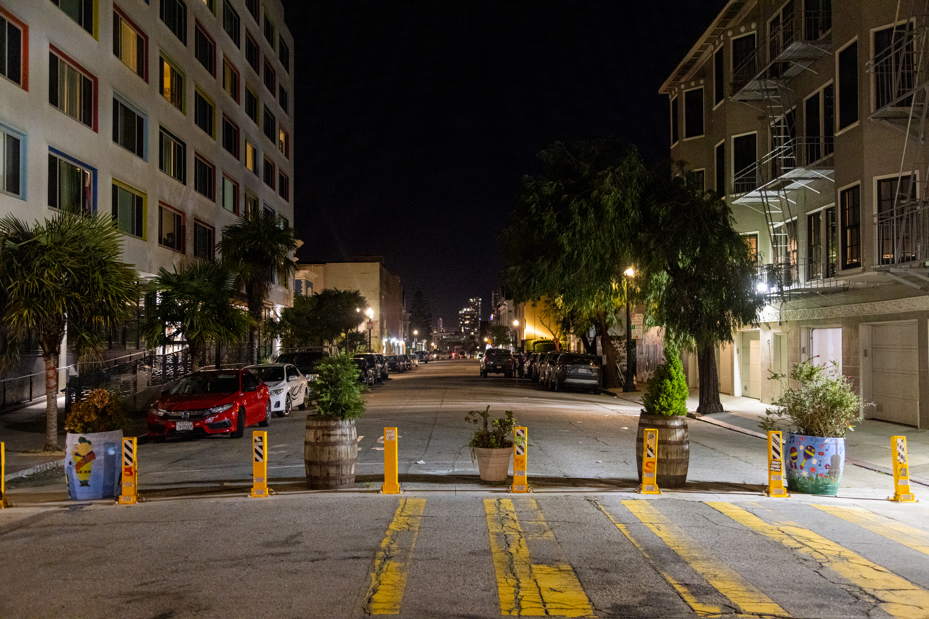 A nighttime street scene with buildings on both sides, parked cars, and planters with plants blocking the street. Yellow and black striped barriers are placed along the road.