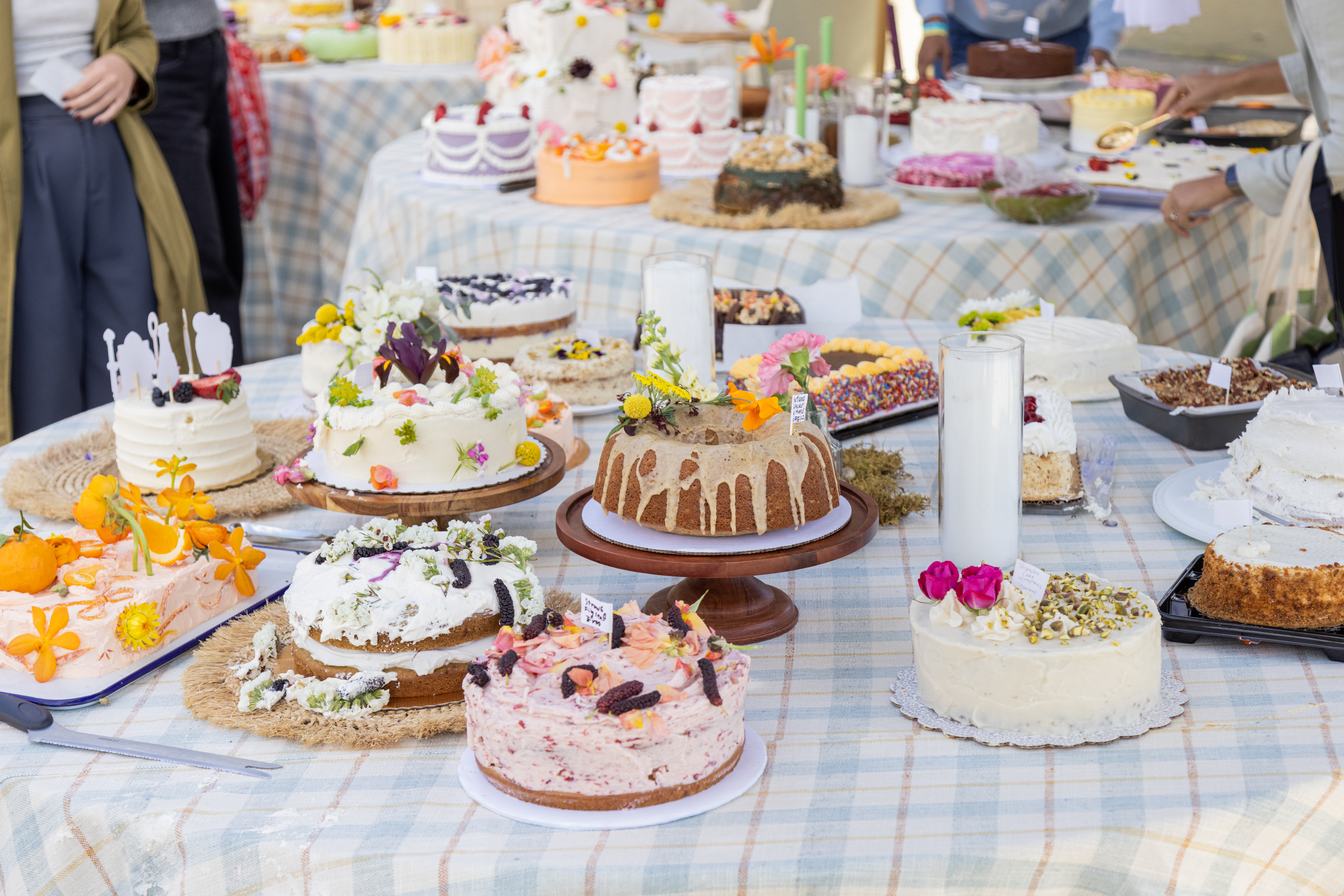 A table filled with various decorated cakes, some topped with flowers, displayed outdoors.