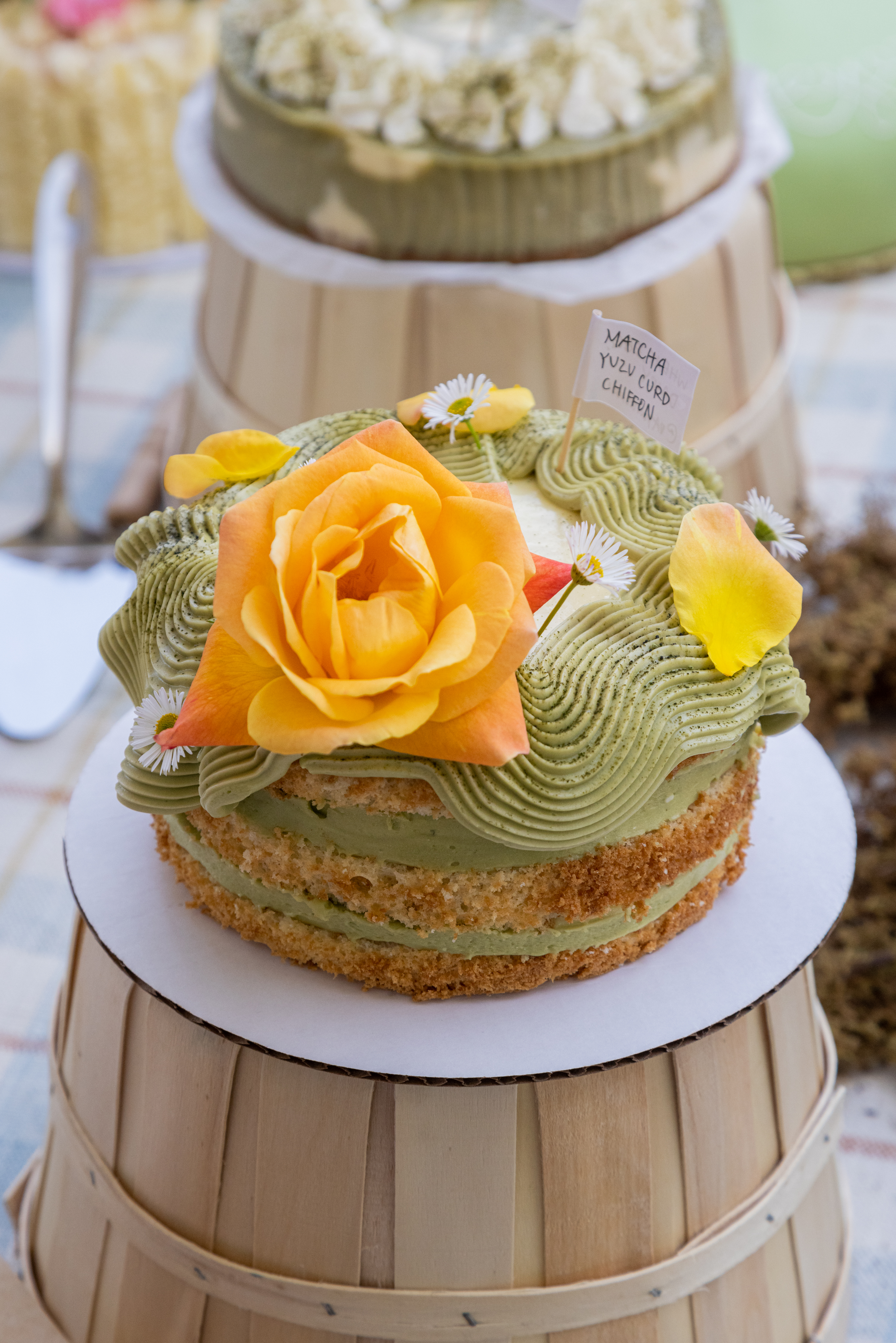 A matcha cake with a vibrant orange rose on top sits on a wooden stand, surrounded by other pastries.