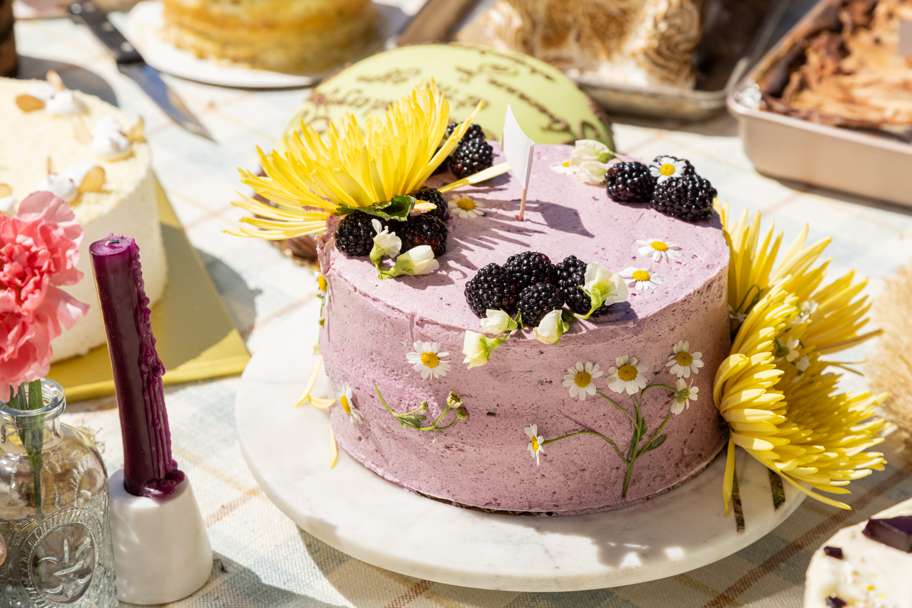 A purple cake adorned with blackberries, chamomile flowers, next to bright yellow blooms on a sunny table.