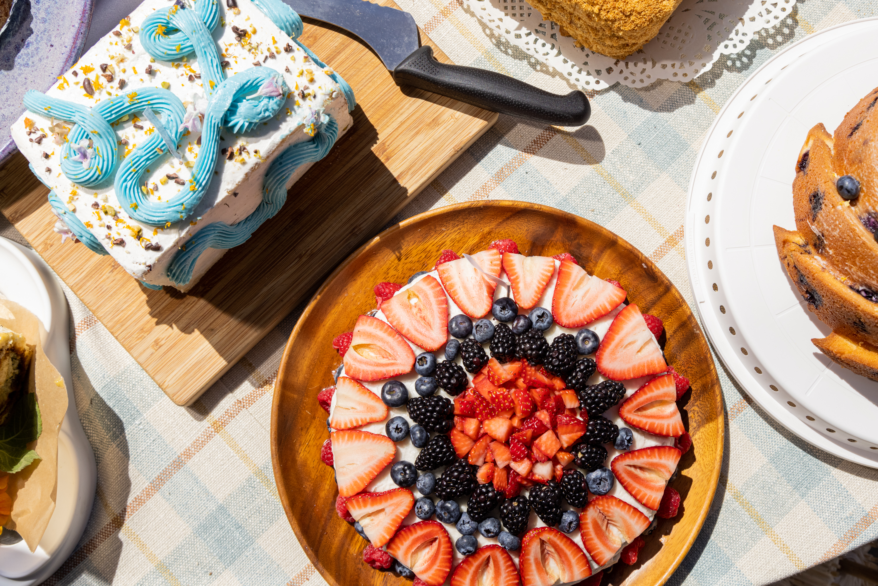 An array of desserts on a sunny table with a fruit tart, a blue swirl-topped cake, and a blueberry cake.