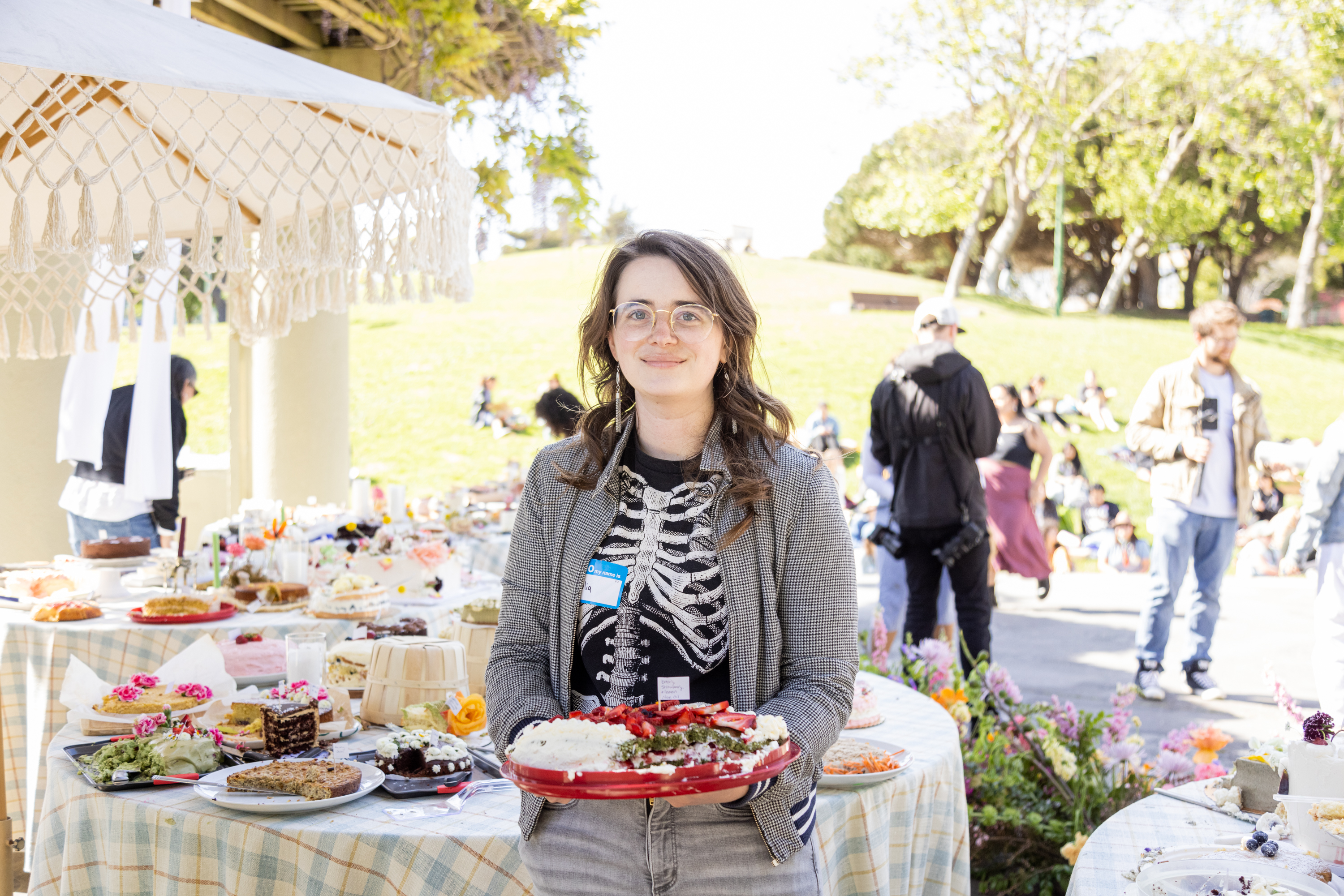A woman holding a dish stands in front of a table laden with food at an outdoor event.