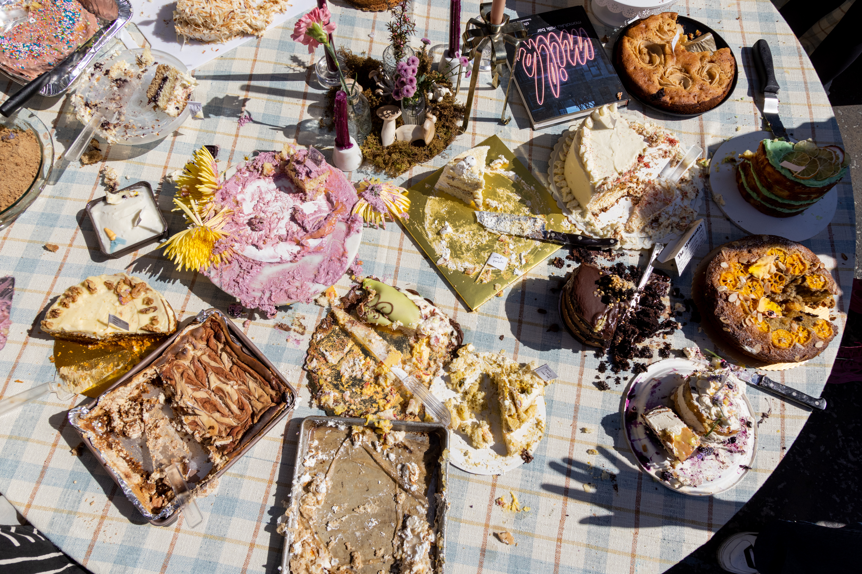 A table with various half-eaten cakes and pies, adorned with flowers and a &quot;Yum&quot; sign, in bright daylight.