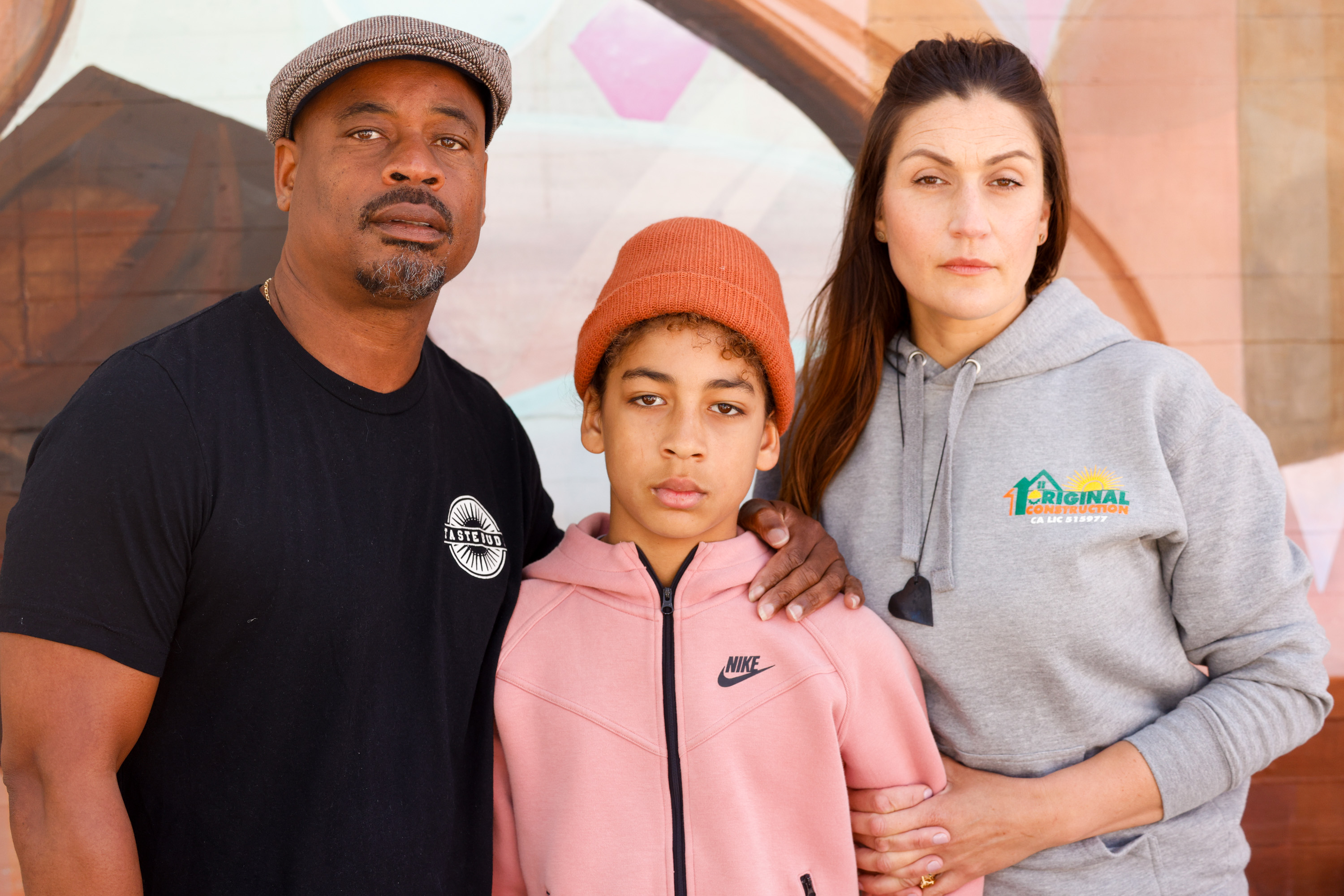 A family of three, with the parents flanking a teenager, stands together in front of a mural.