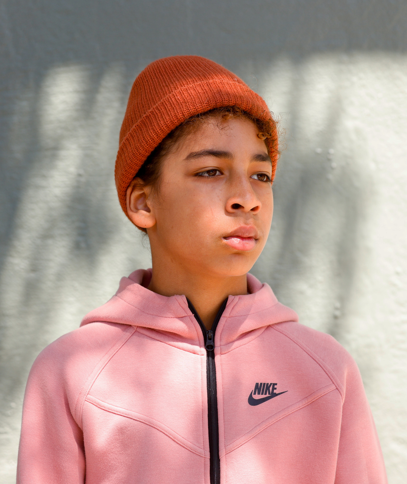 A young person wearing a pink jacket and an orange beanie stands in front of a grey wall, looking to the side.