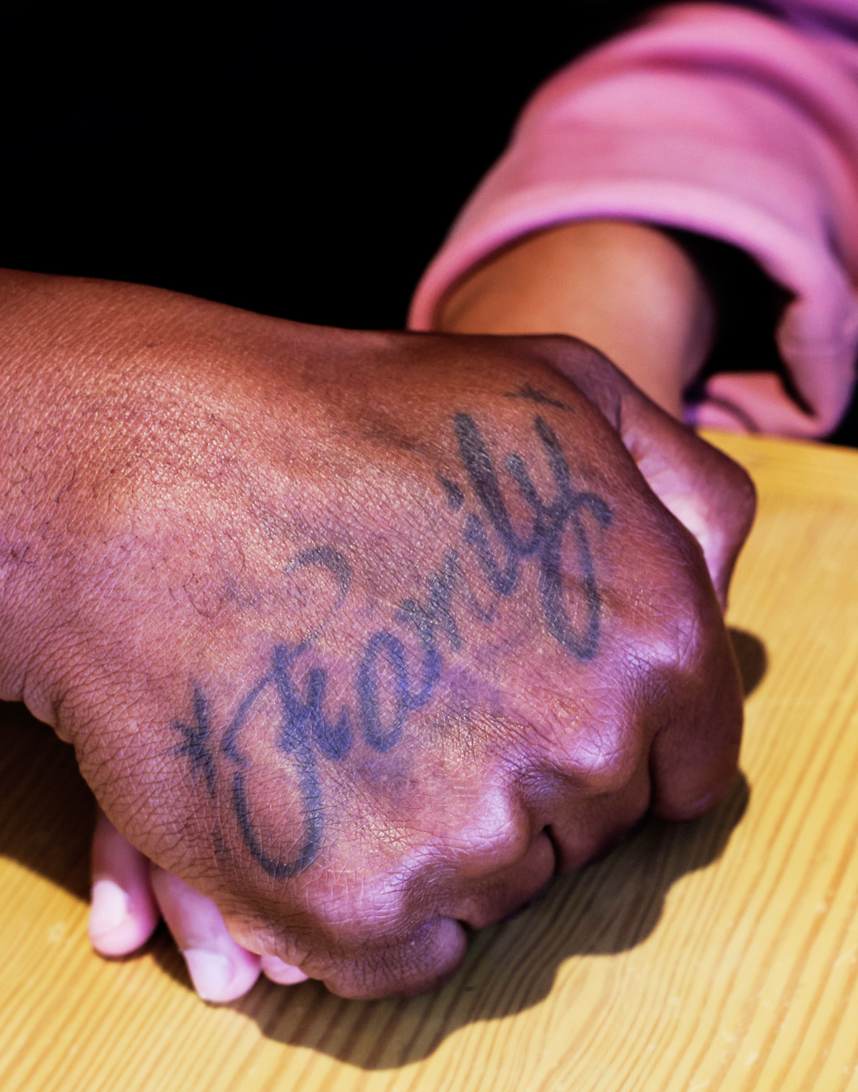 A hand with a &quot;Family&quot; tattoo resting on another hand atop a wooden surface.