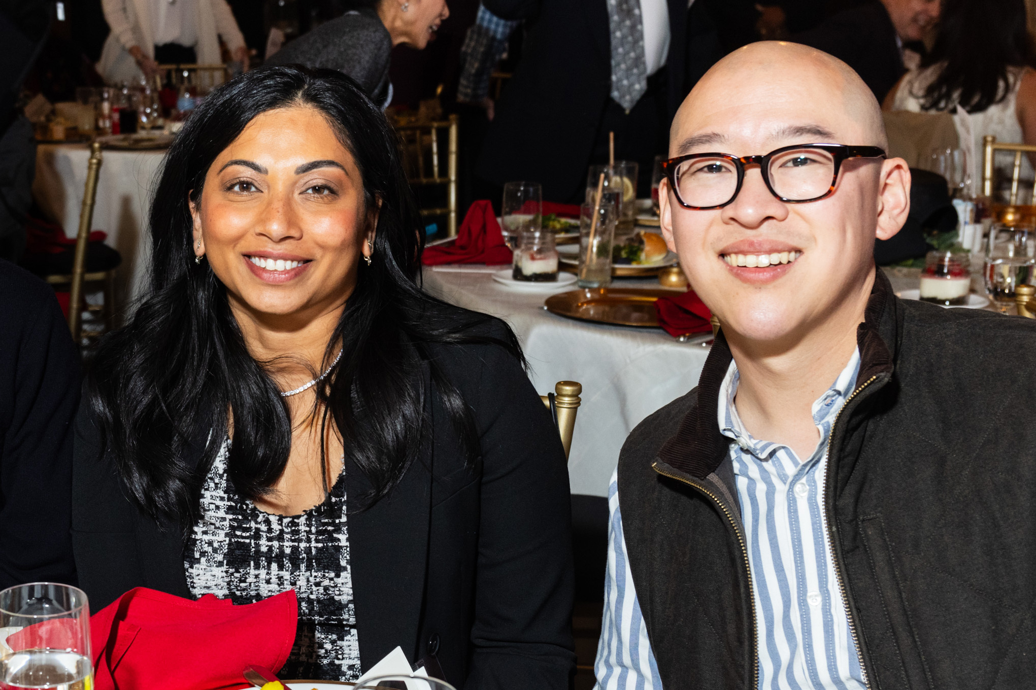 Kanishka Cheng and Jay Cheng both smile while seated at a festive table with dinnerware, both dressed semi-formally.
