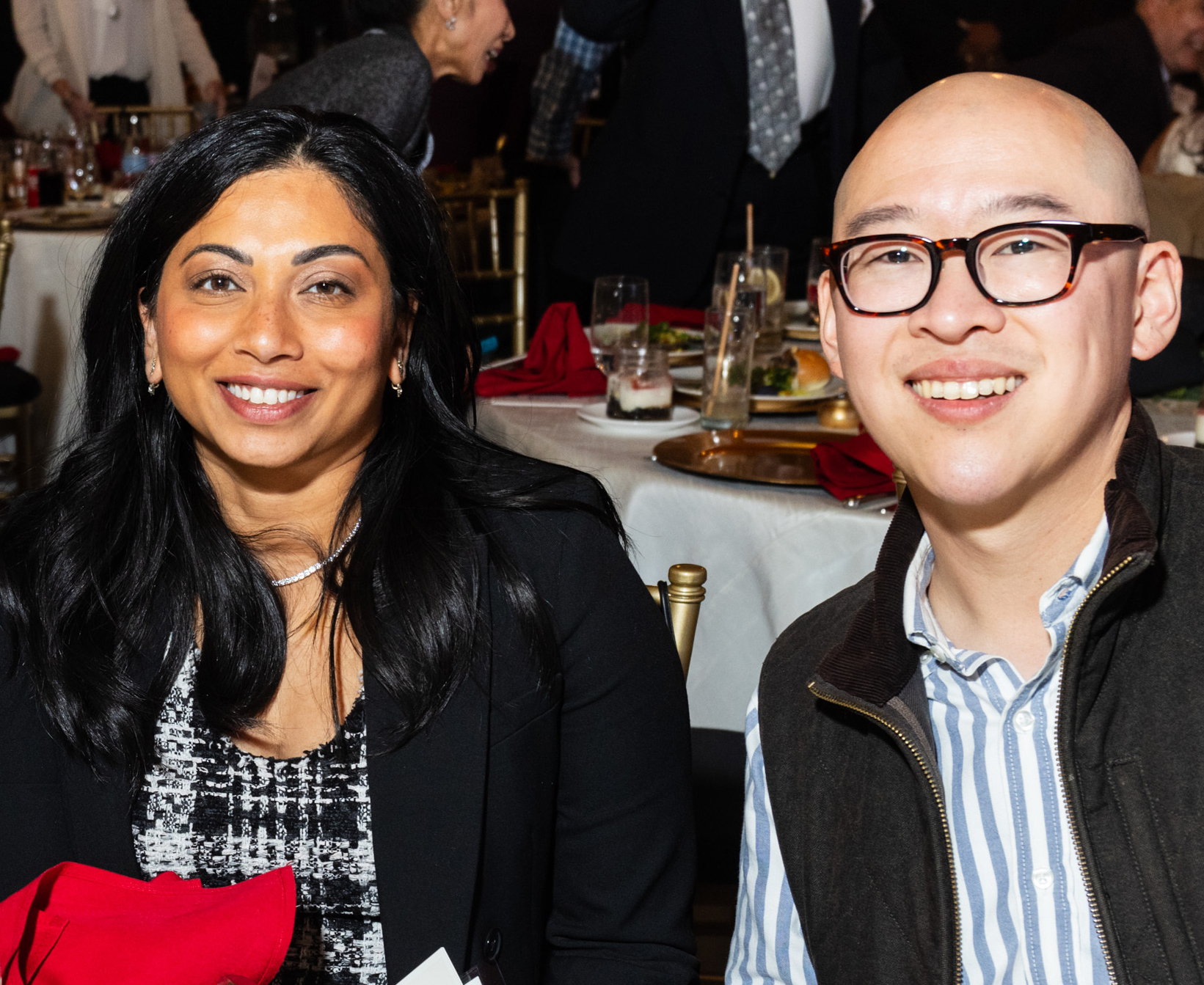 Kanishka Cheng and Jay Cheng smile a table at a social event. She has long hair and he wears glasses.