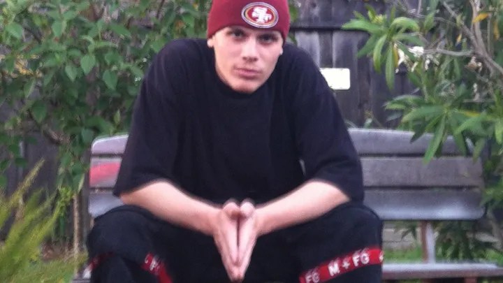 A person is squatting, hands together, wearing a 49ers cap, black shirt, and pants with red accents, in a garden with a bench.