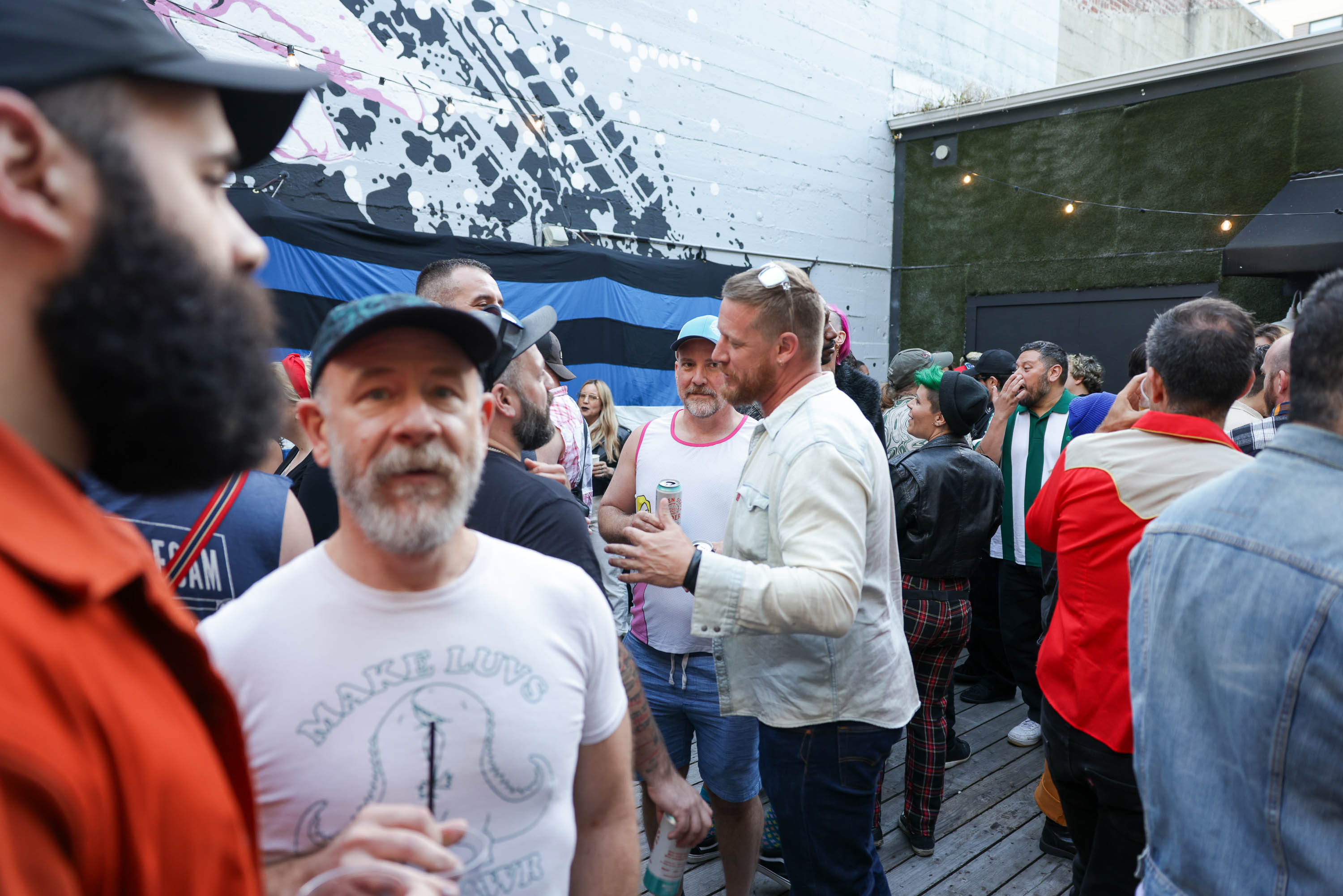 a bearded man in a white shirt and blue cap holds a drink on a crowded patio