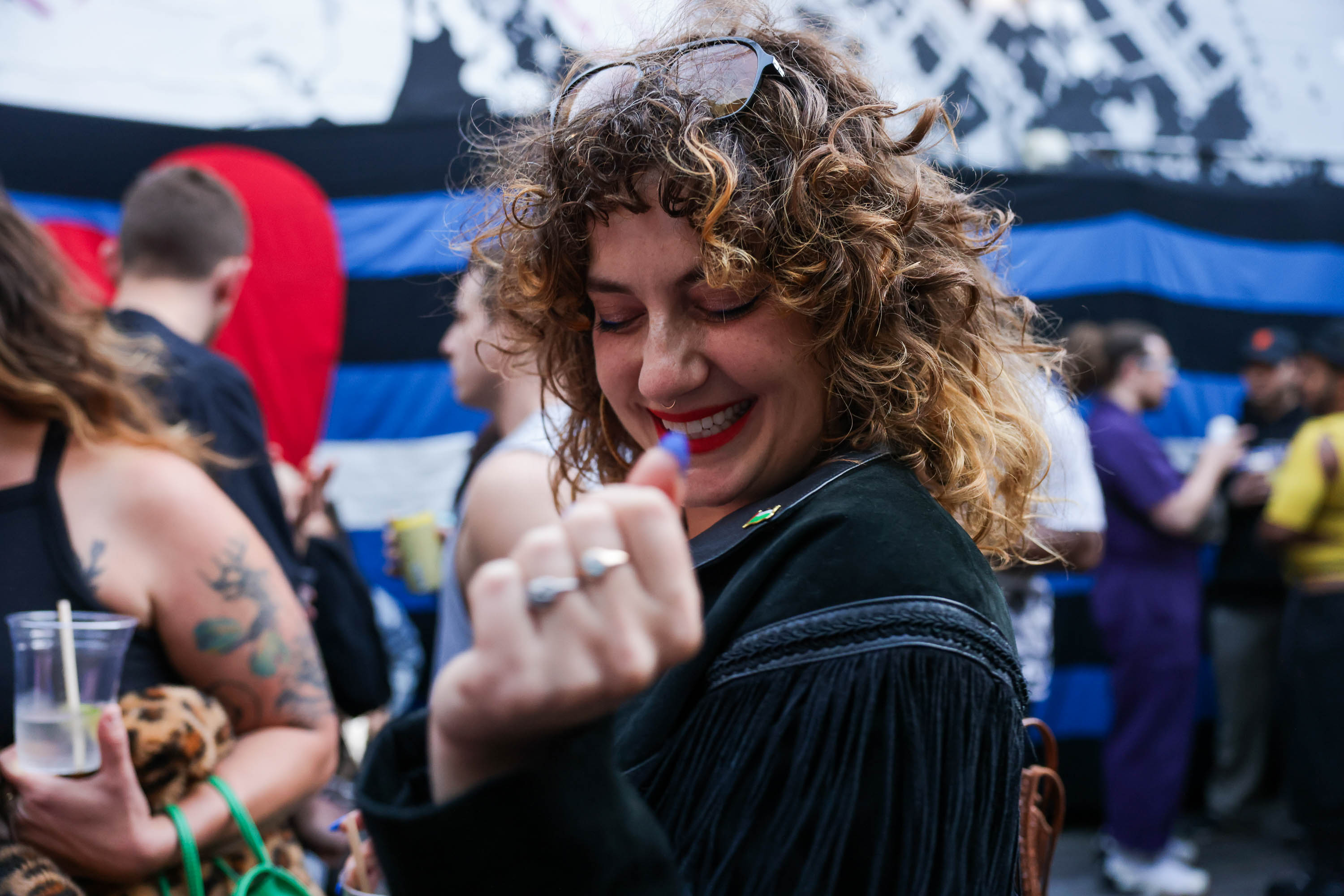 A smiling woman with curly hair, holding a clear cup, enjoys a bustling outdoor event.