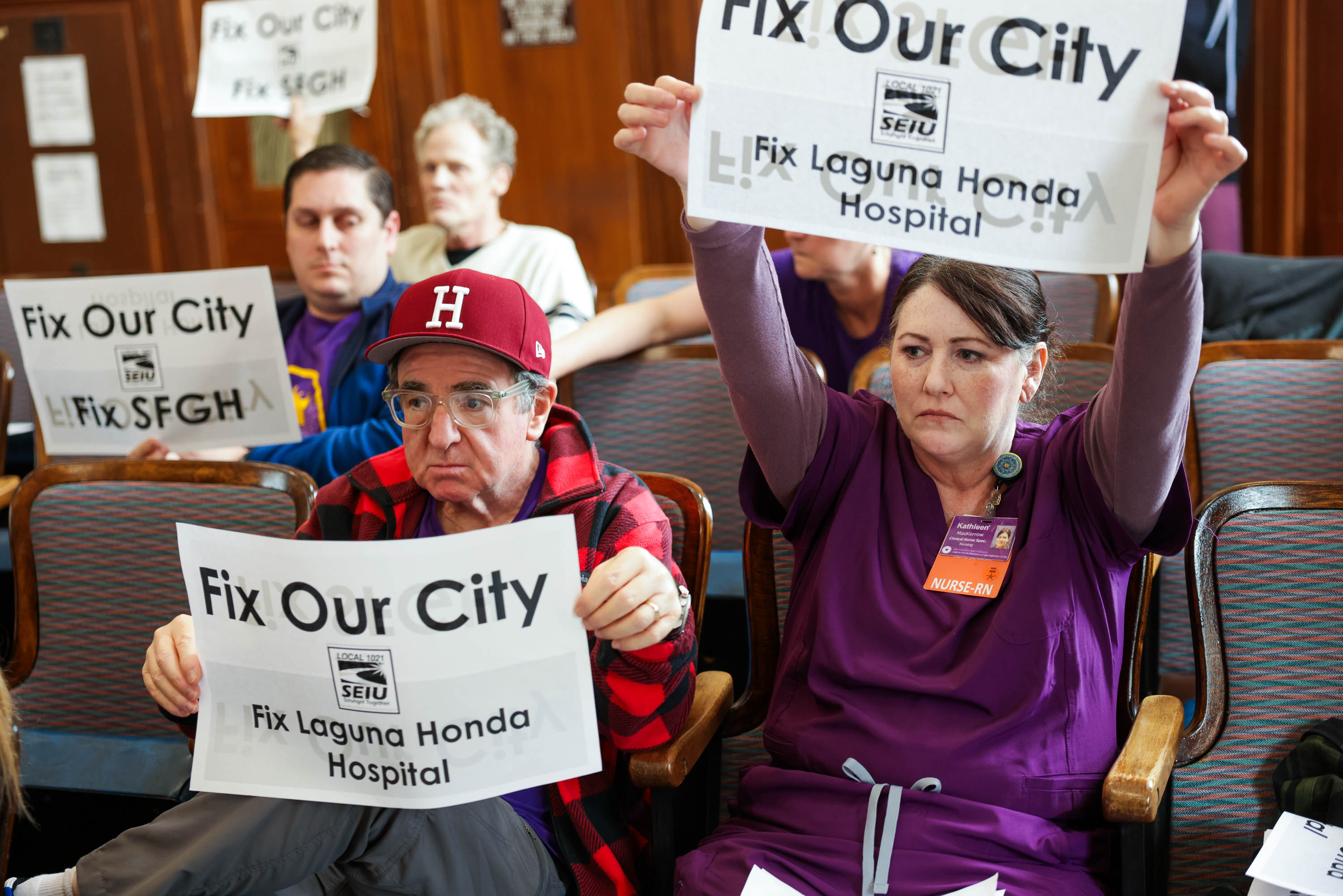 A group of people in a room hold signs saying &quot;Fix our city, Fix Laguna Honda Hospital.&quot; They appear serious and are seated, some wearing work uniforms.