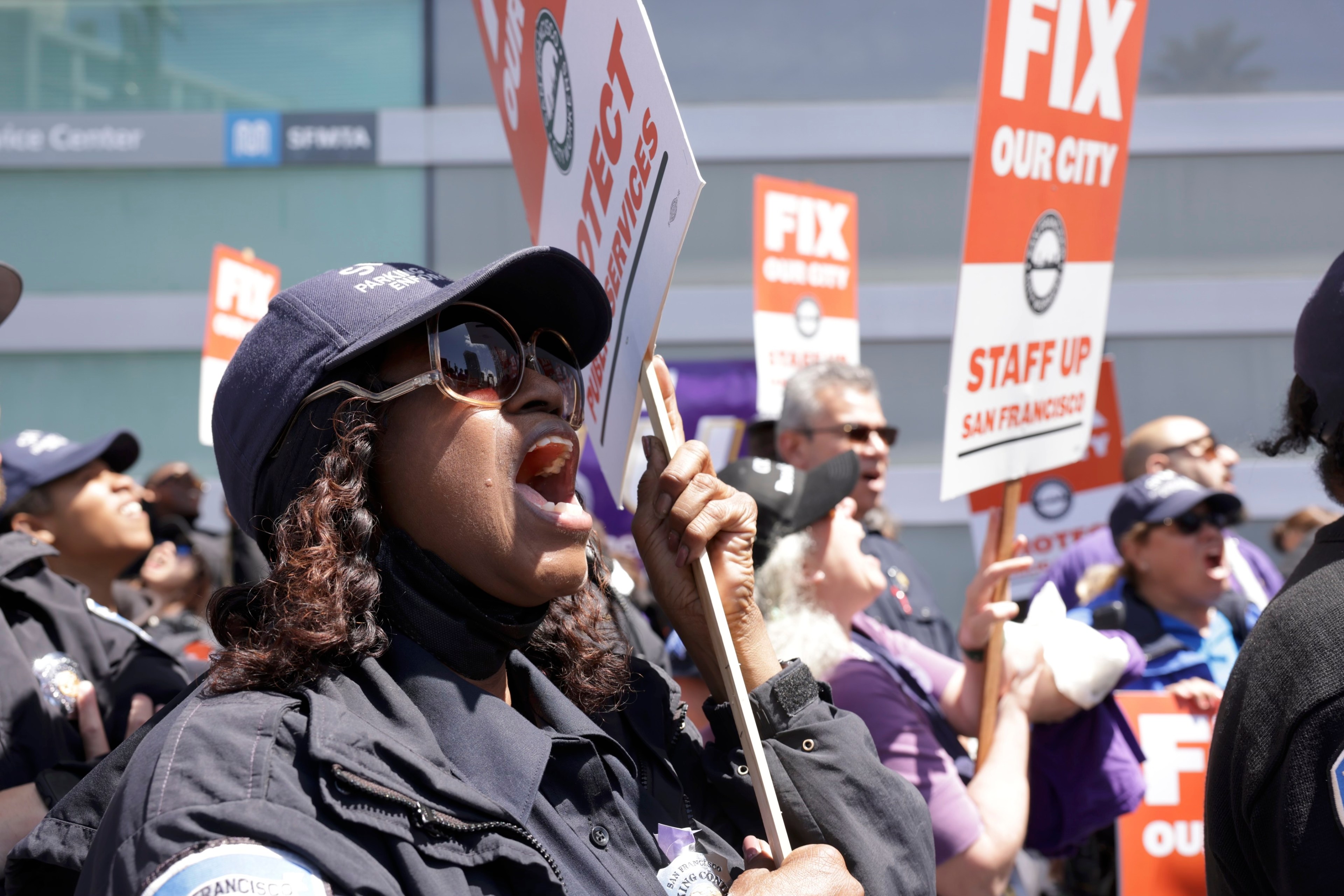 A group of people protesting, holding signs like "FIX OUR CITY." A woman in the foreground shouts, wearing sunglasses and a cap.