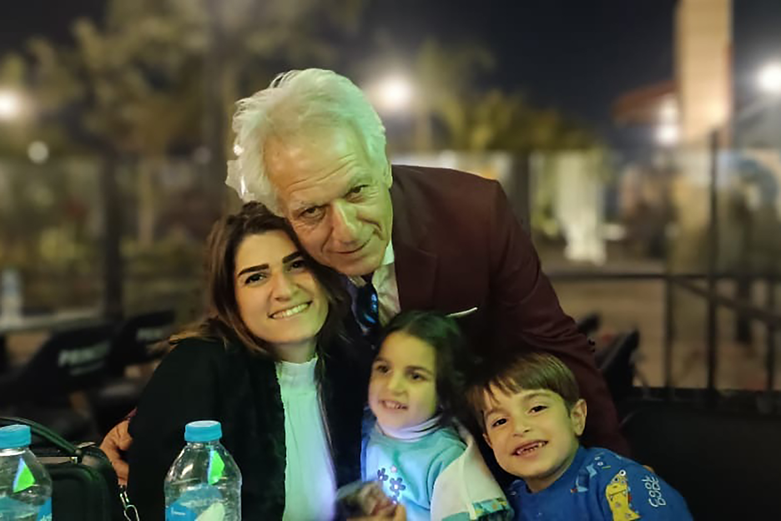 An older man in a suit hugs two smiling children and a woman outdoors at night.
