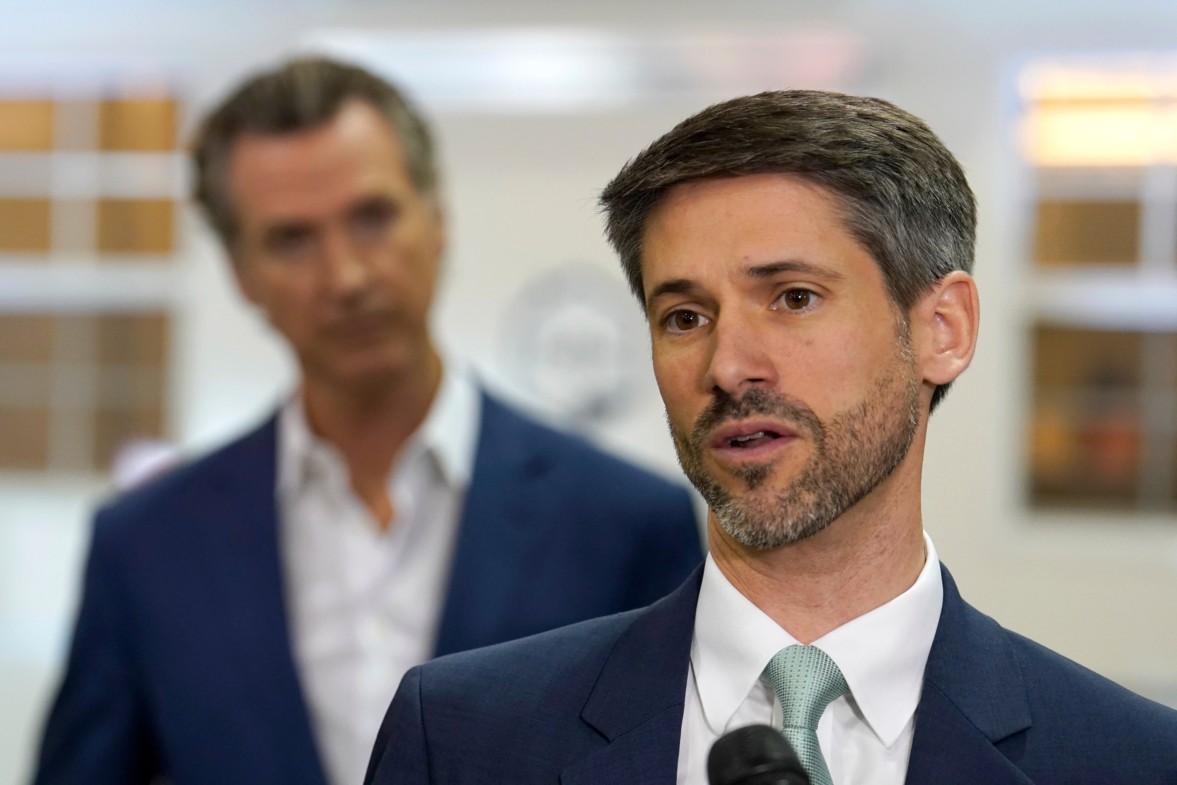 Two men in suits, one speaking into a microphone, the other slightly out of focus in the background.