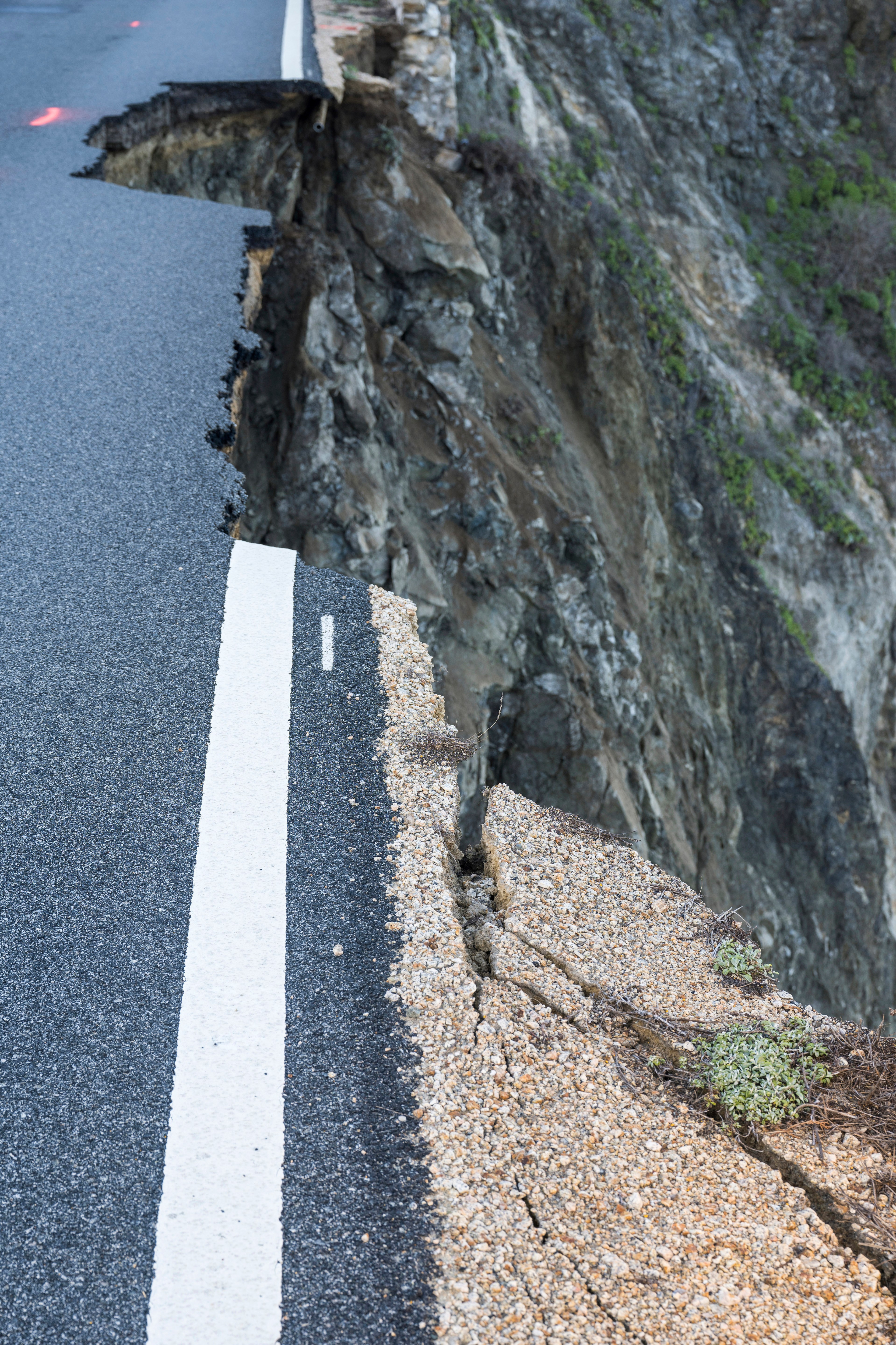 A road abruptly ends at a collapsed cliff edge, with barriers partially visible.